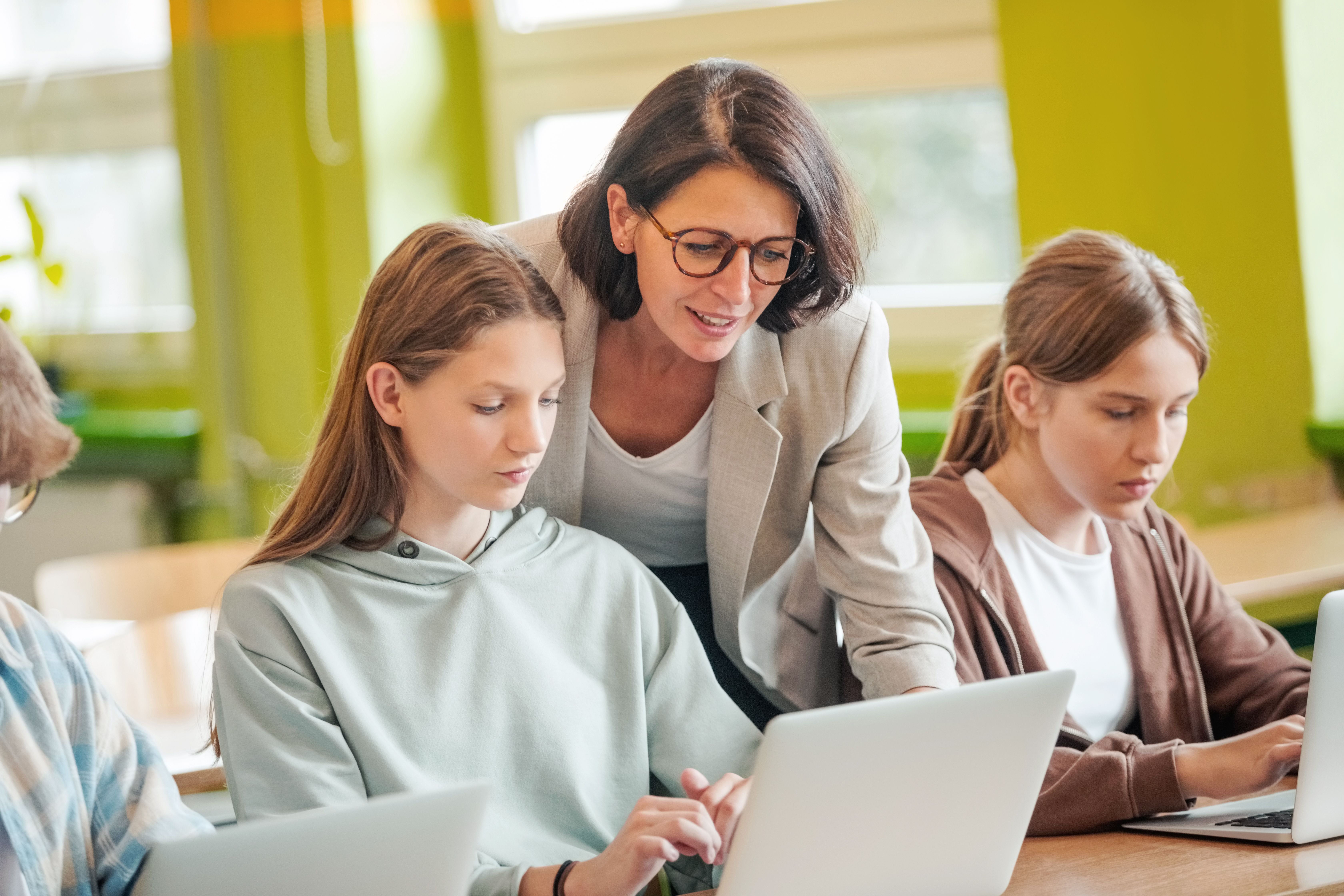 Lehrerin hilft zwei Schülerinnen im Klassenzimmer, die an Laptops arbeiten.
