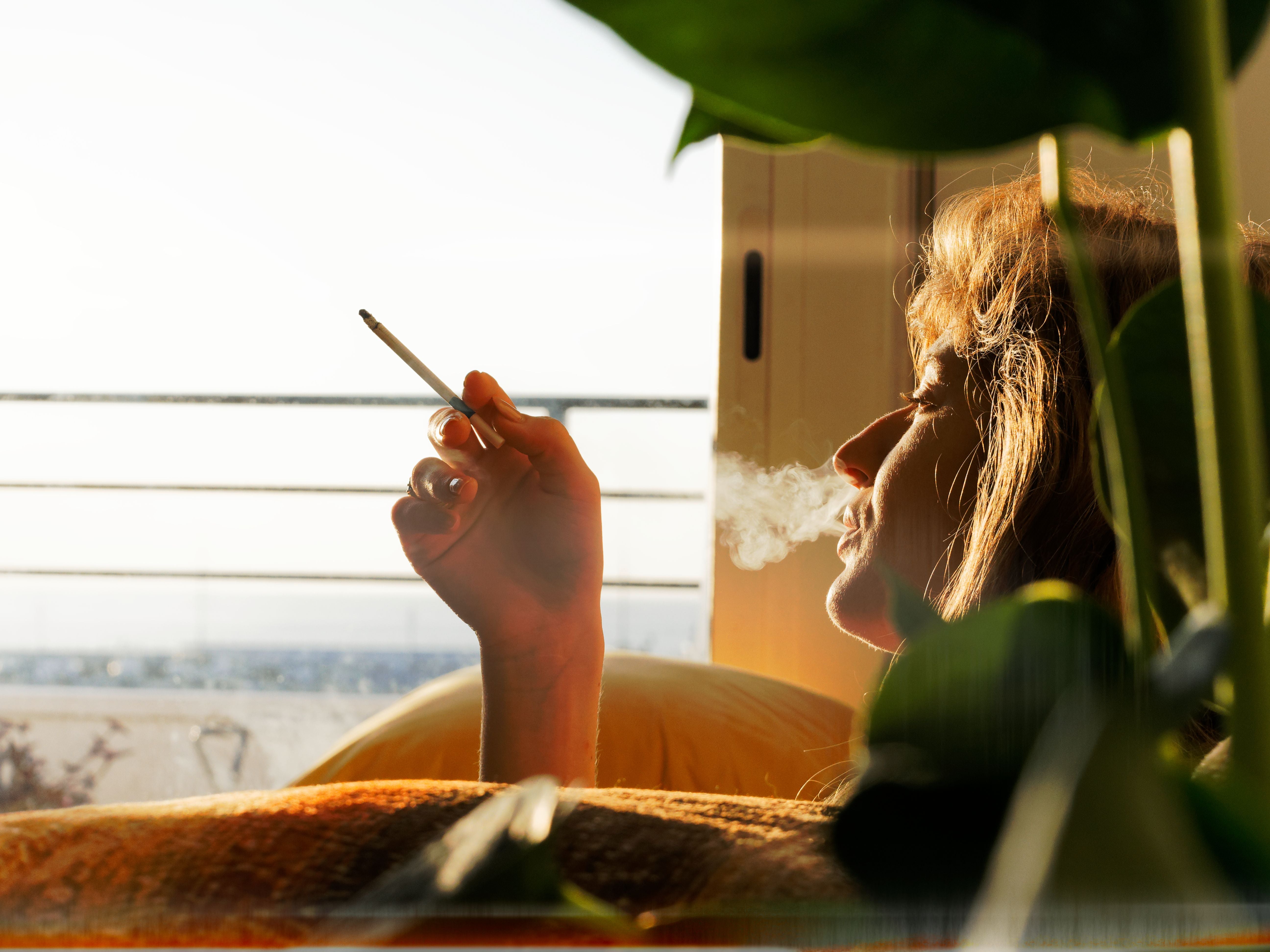 Blonde Frau raucht eine Zigarette und bläst Rauch aus, Sonnenlicht scheint durchs Fenster.