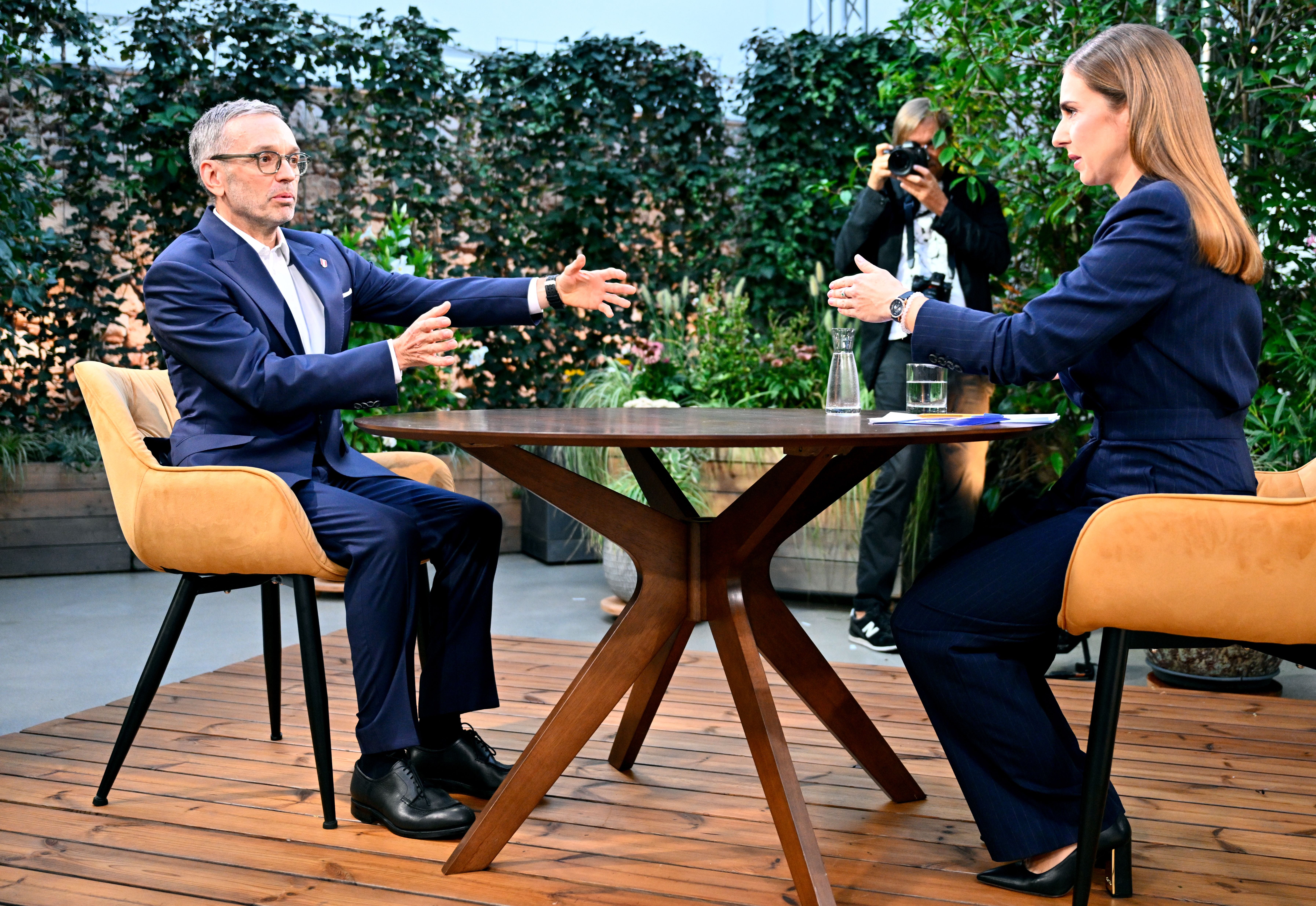 Ein Mann und eine Frau sitzen sich bei einem Fernsehinterview im Freien an einem Tisch gegenüber.