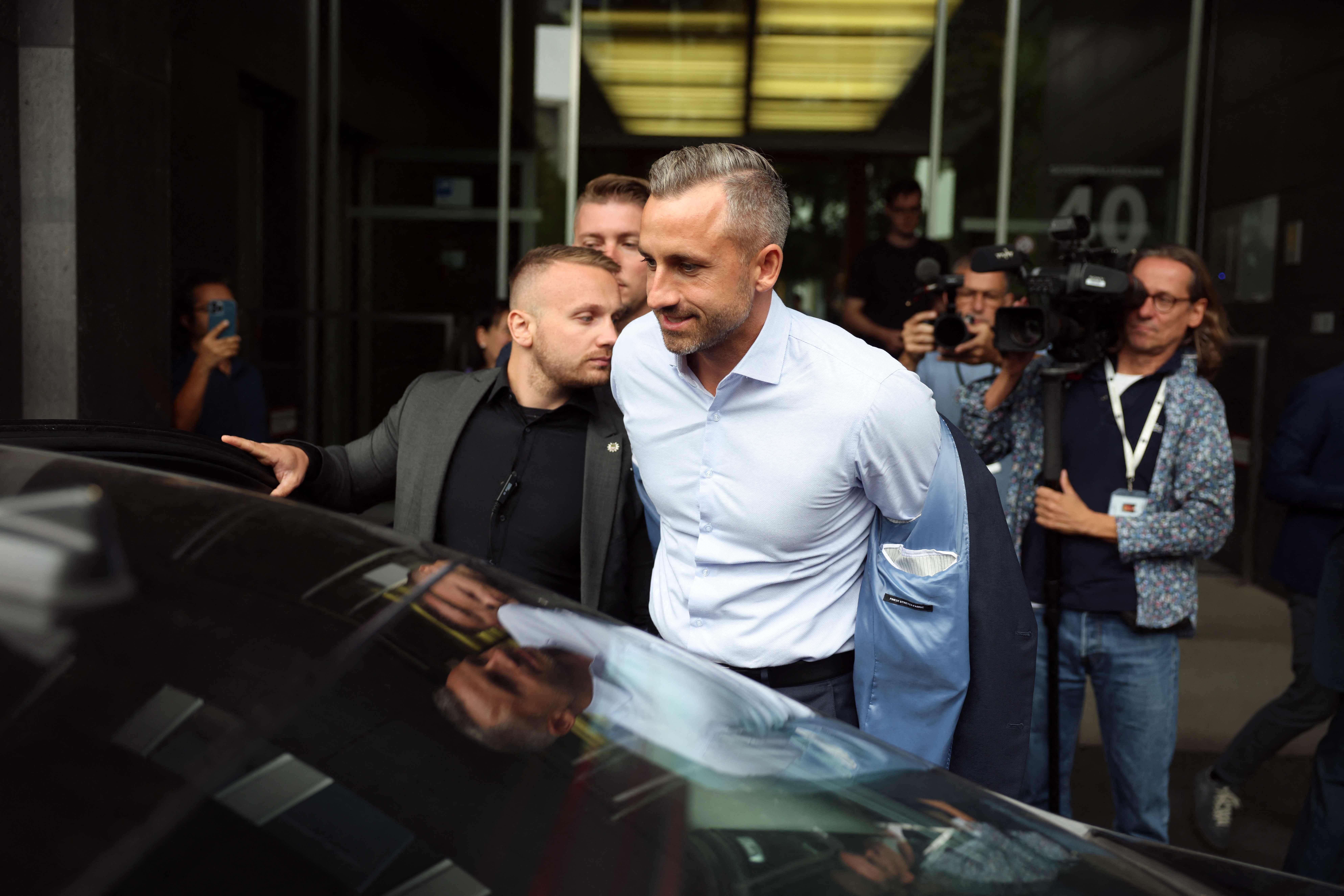 Ein Mann zieht sein Sakko aus und steigt vor Journalisten und Kameraleuten in ein Auto ein.