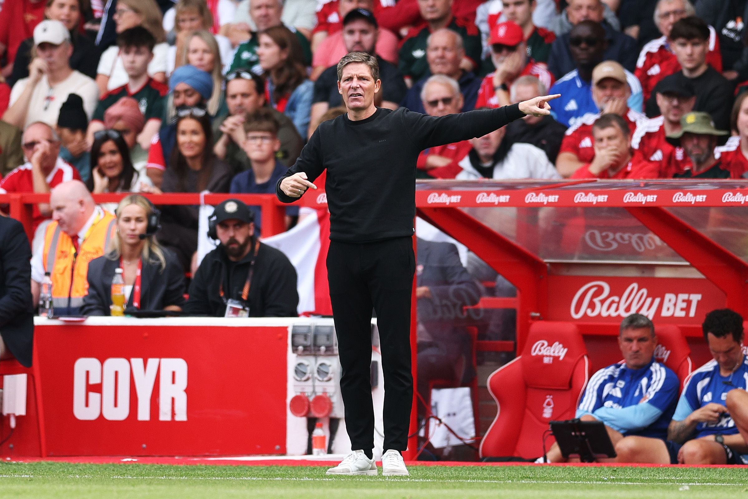 Ein Trainer gibt Anweisungen am Spielfeldrand, Zuschauer sitzen im Hintergrund.