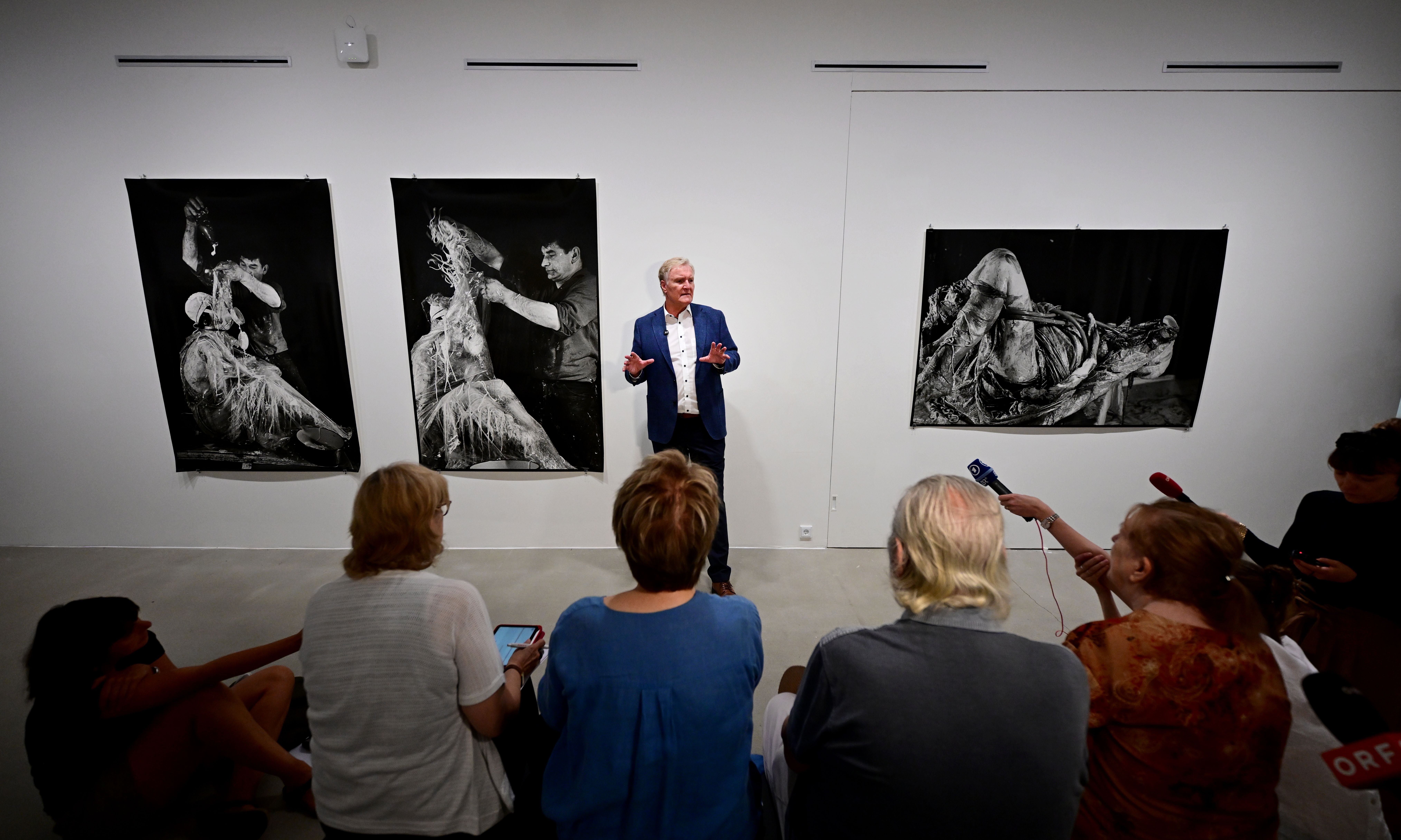 Ein Mann spricht vor einer Gruppe in einer Galerie mit drei schwarz-weißen Kunstdrucken an der Wand.