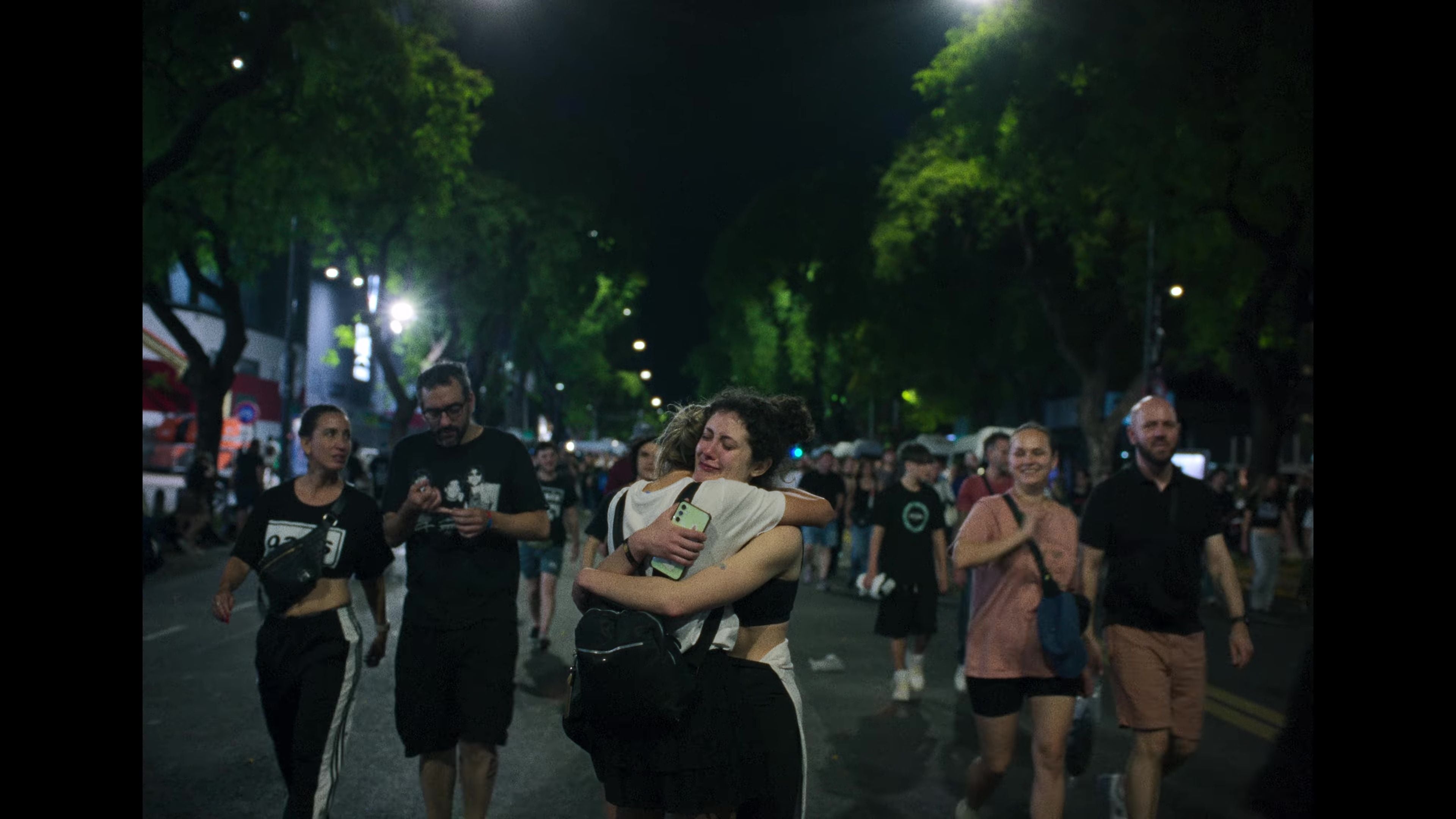 Zwei Frauen umarmen sich emotional auf einer nächtlichen Straße, umgeben von Menschenmengen.