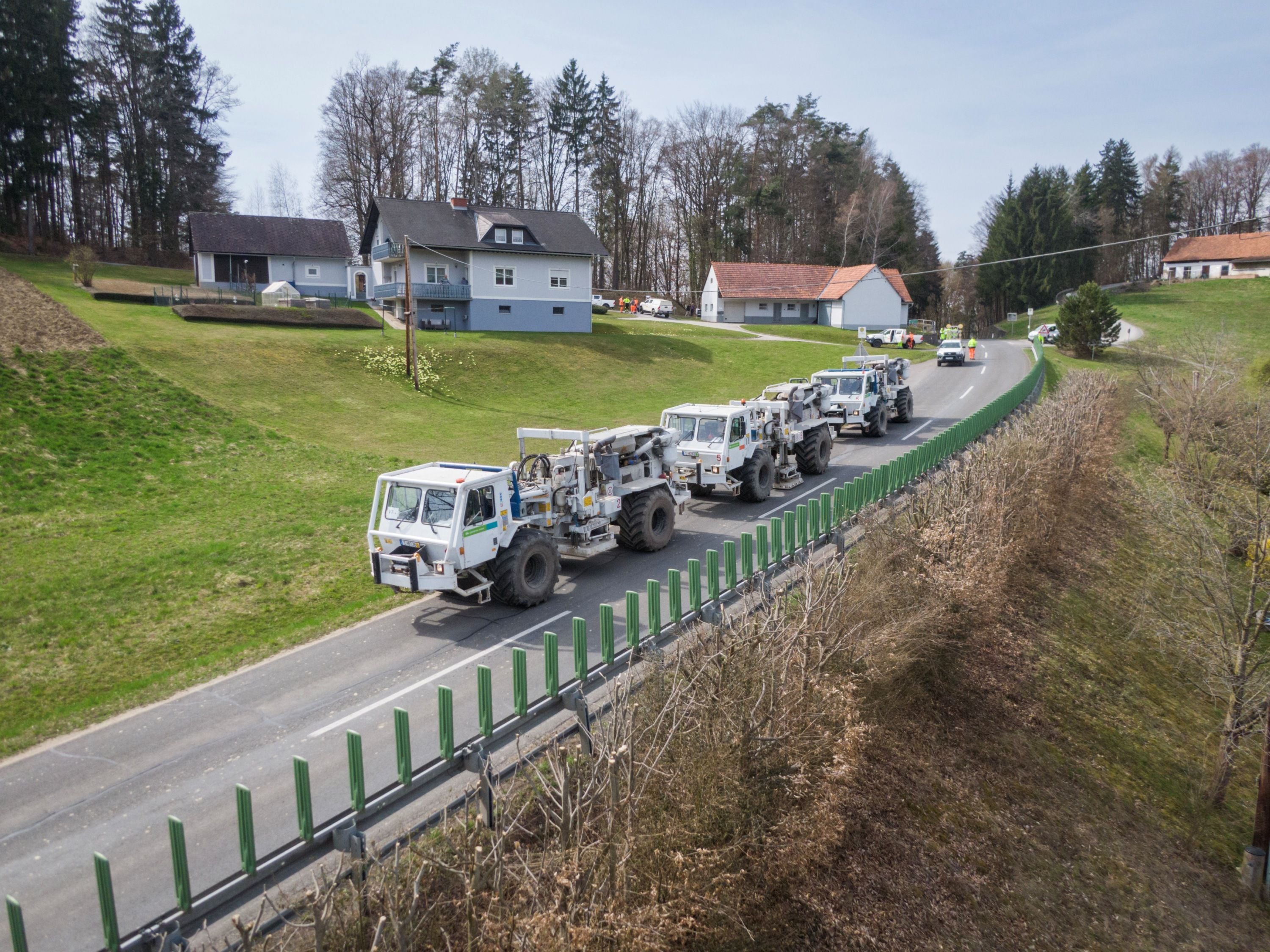 Mehrere Vibro-Trucks fahren in Kolonne auf einer Landstraße durch eine ländliche Gegend.