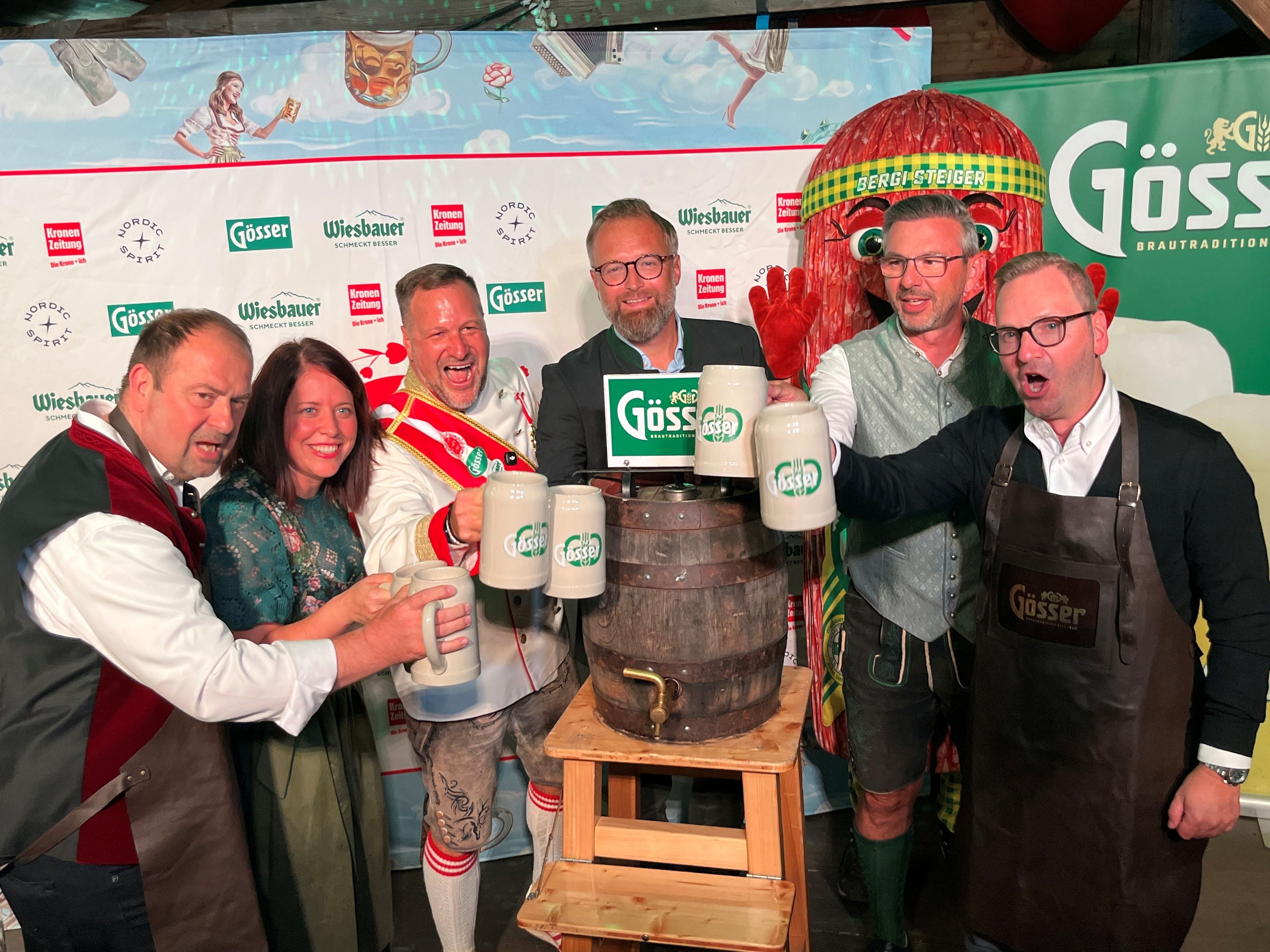 Sechs Menschen stoßen auf der Kaiserwiesn fröhlich mit Bierkrügen um ein Holzfass an.