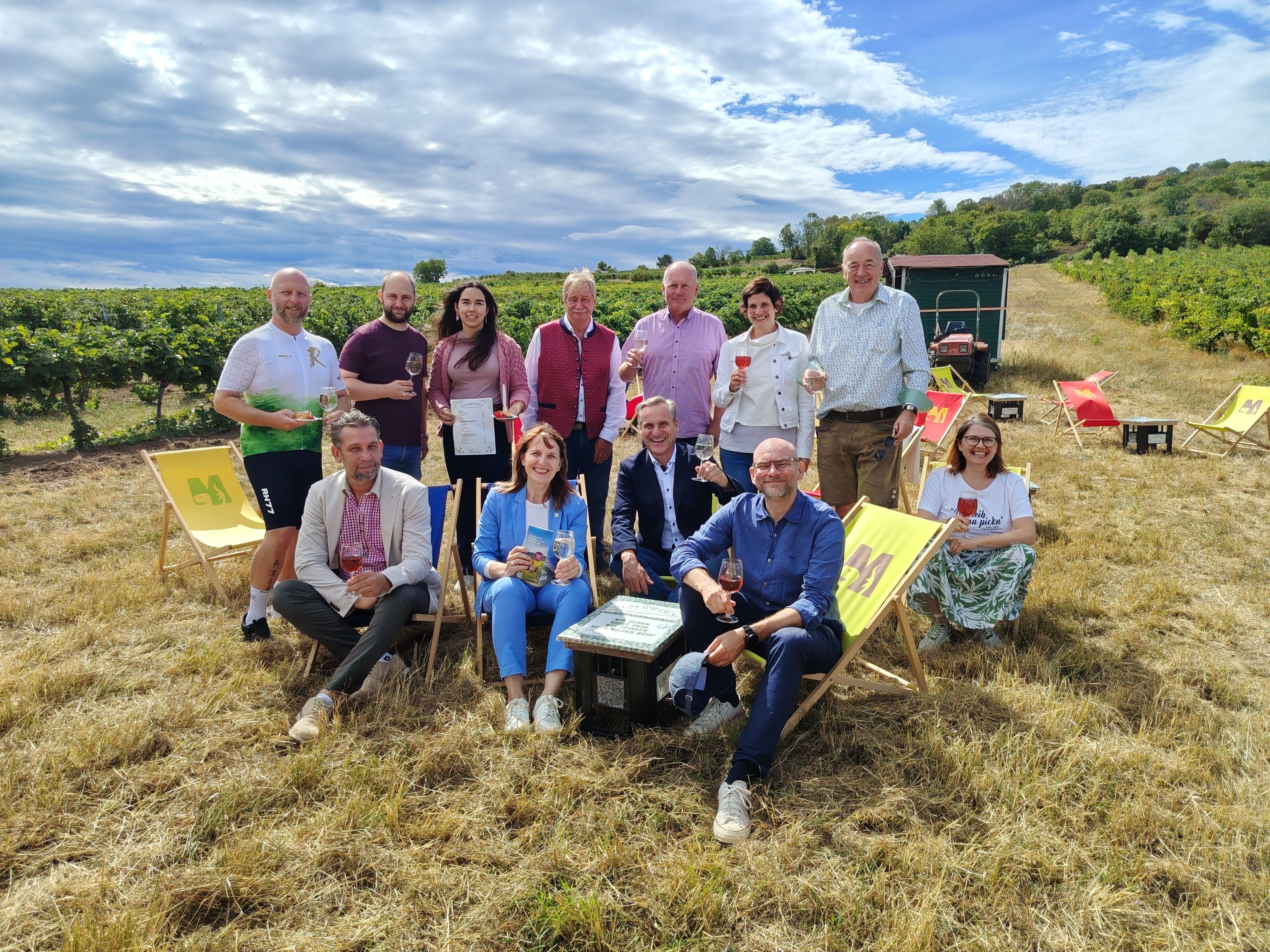 Gruppe von Menschen sitzt und steht mit Weingläsern auf Liegestühlen vor einem Weinberg im Freien.