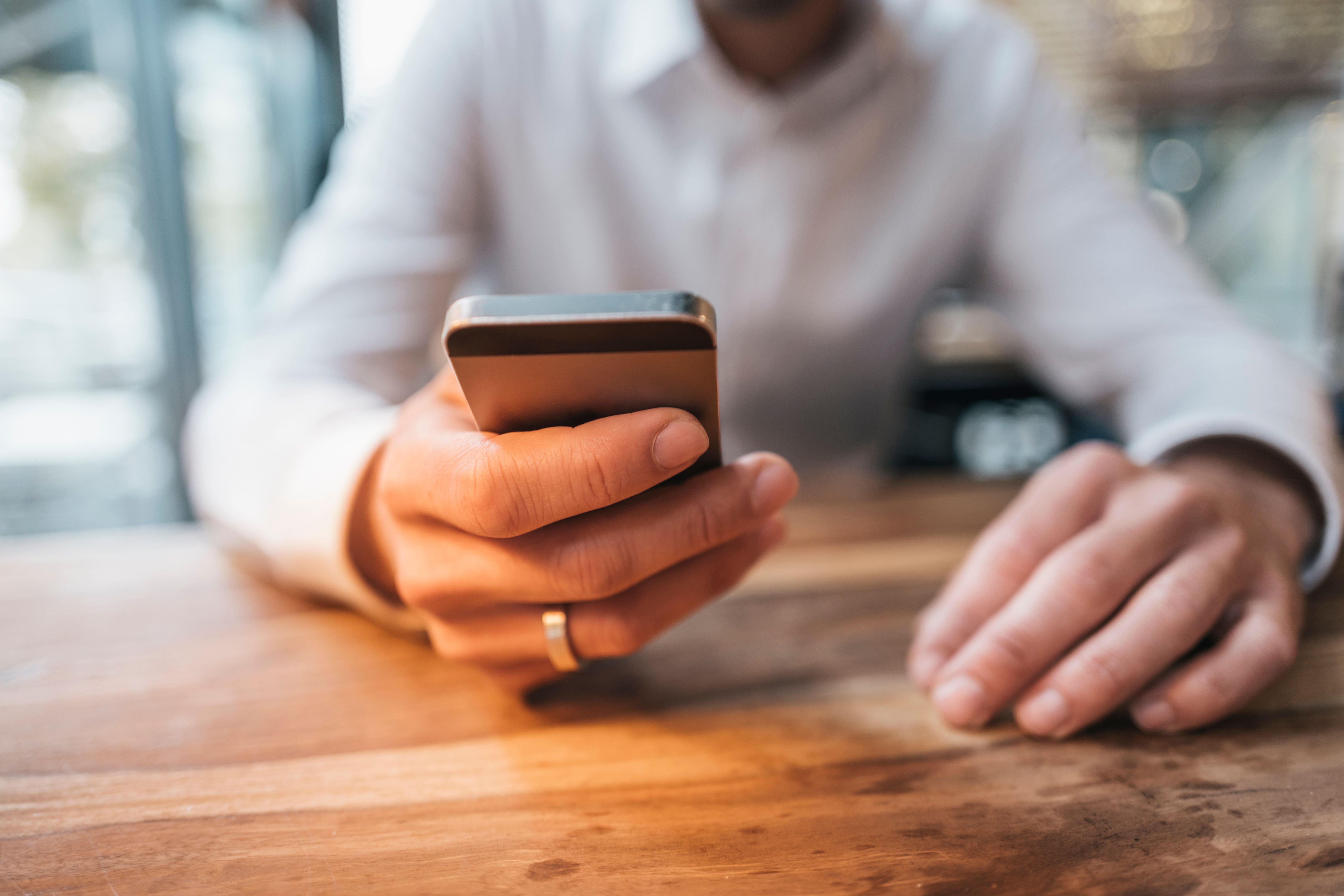 Mann in Hemd hält ein Smartphone über einem Holztisch und schaut darauf.