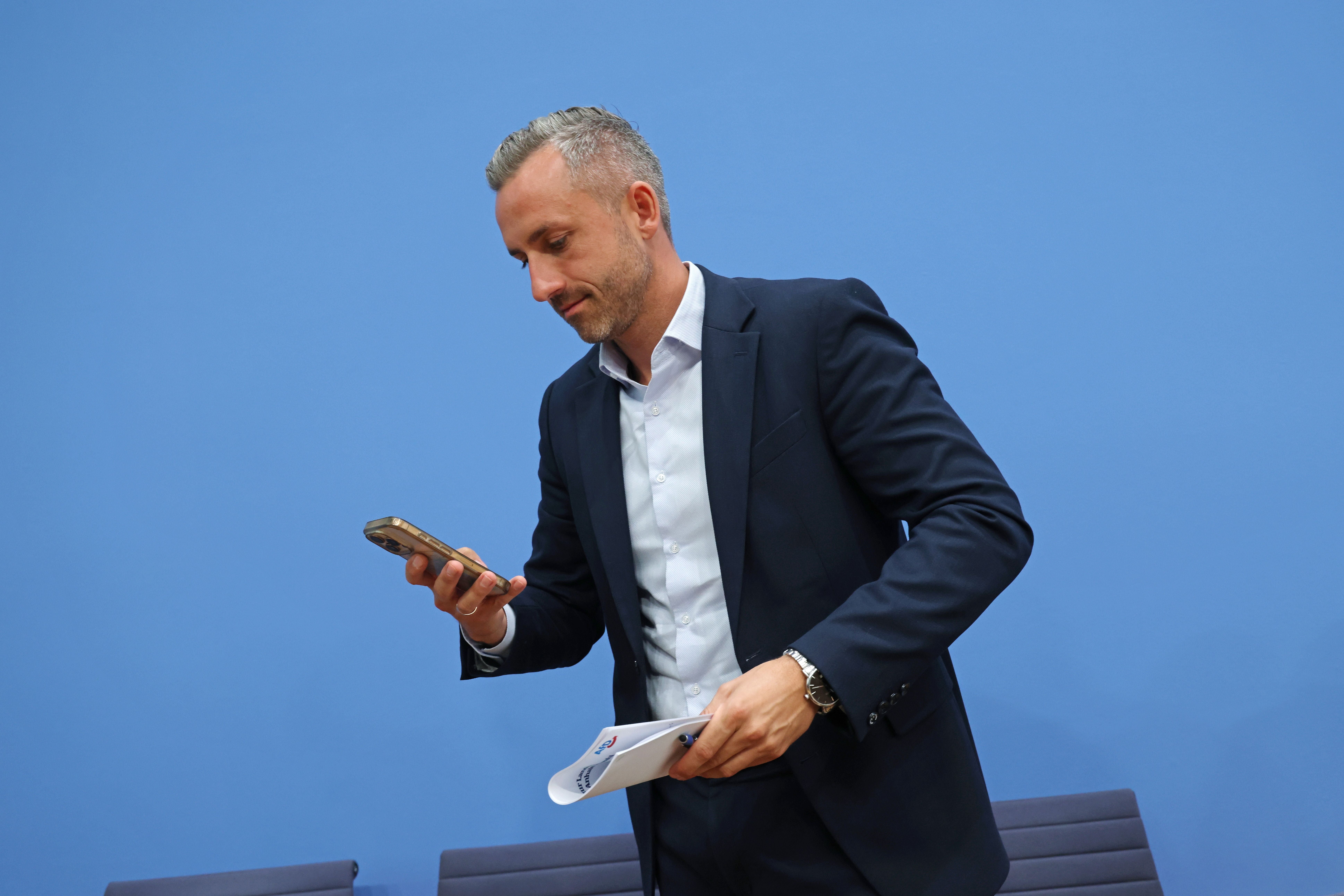 Ein Mann im Anzug schaut auf sein Smartphone und hält Unterlagen vor einer blauen Wand.