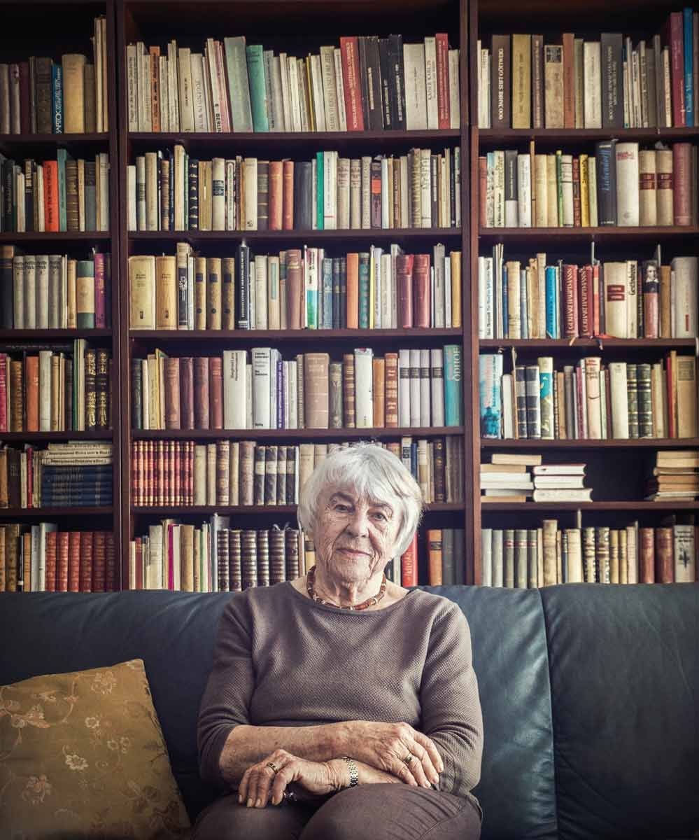 Ältere Frau sitzt auf einem Sofa vor einem großen Bücherregal voller Bücher.
