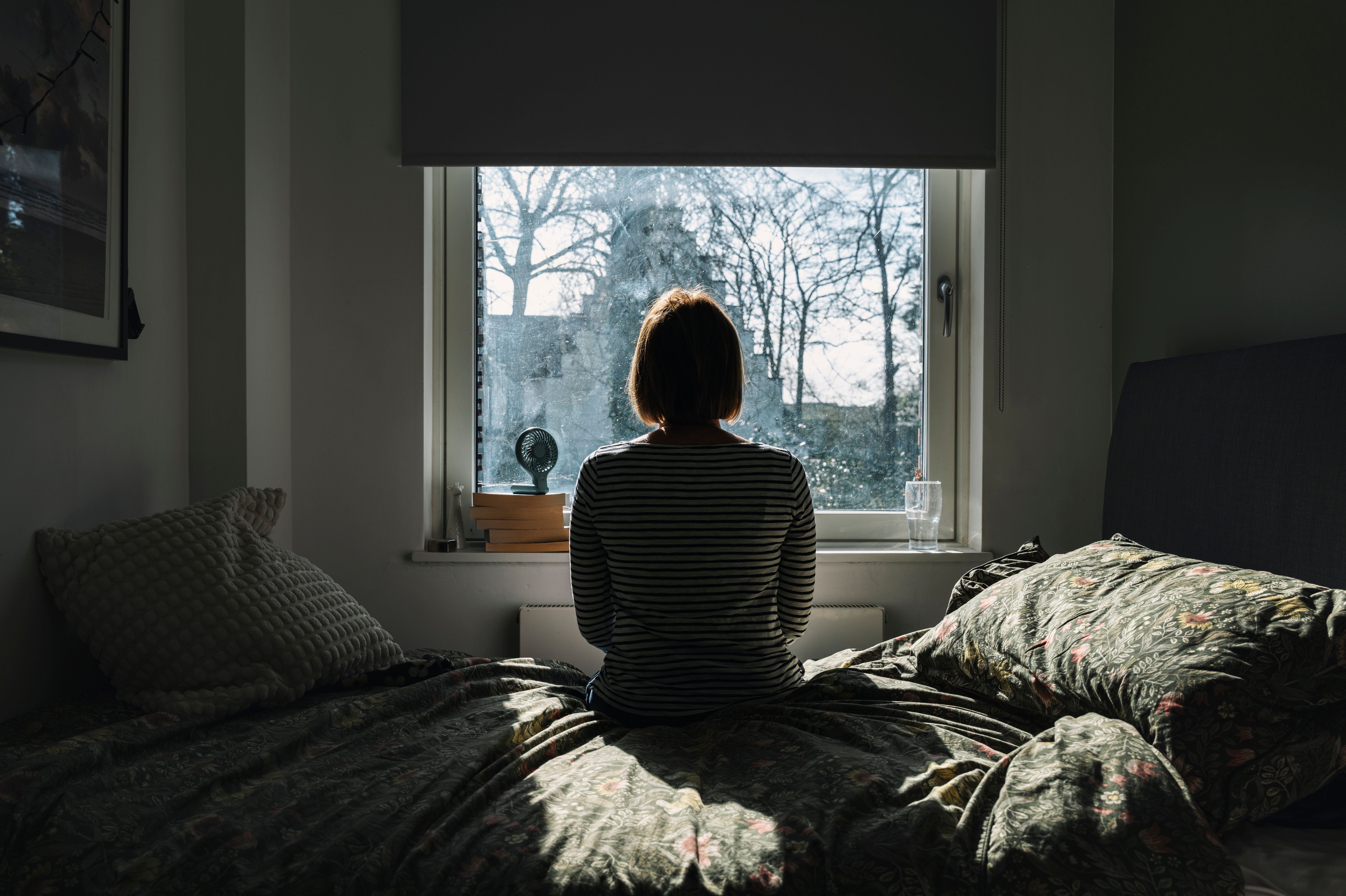 Eine Person sitzt auf einem Bett und schaut aus einem Fenster in einen sonnigen Garten.