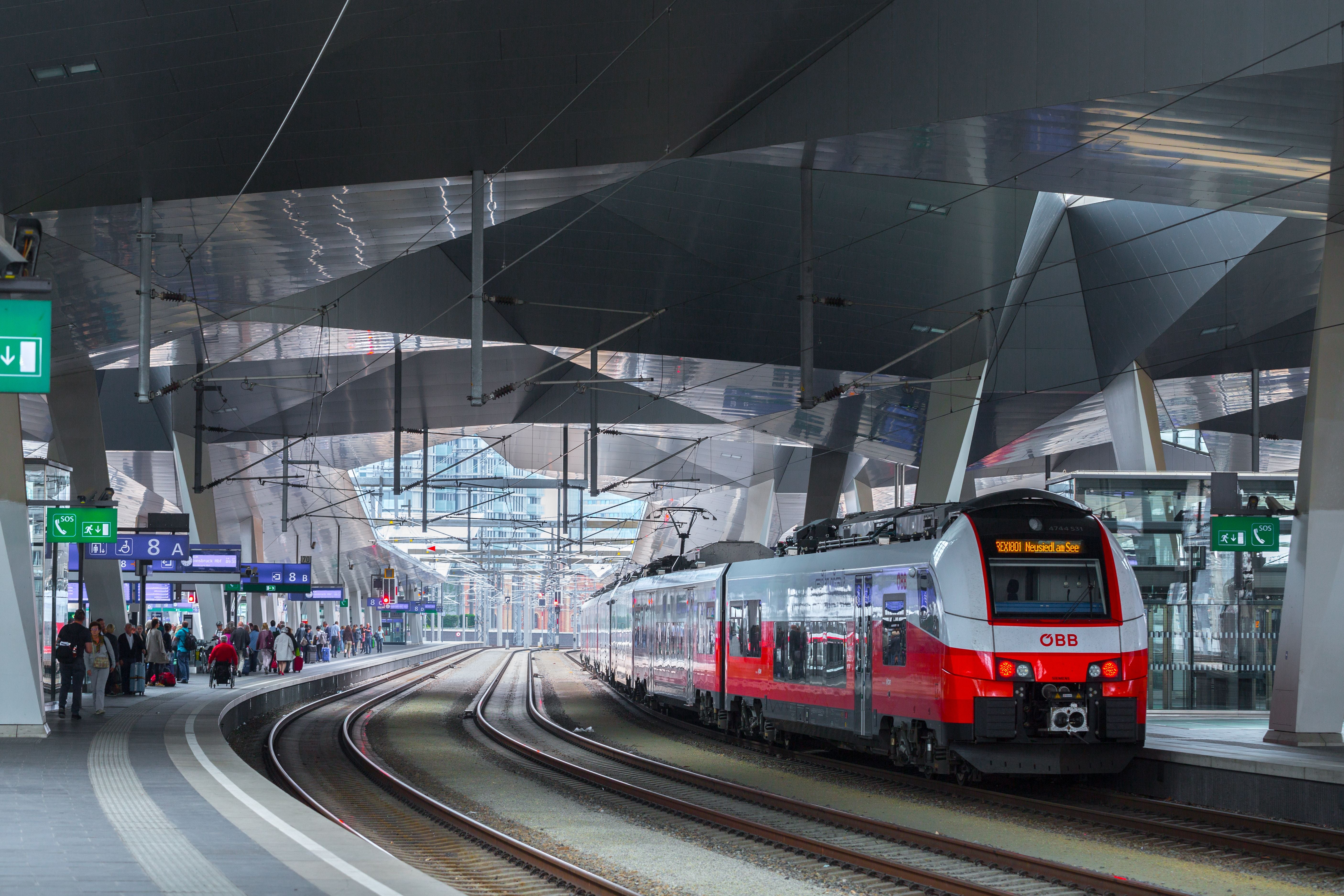 Moderner Bahnhof mit einem ÖBB-Zug und wartenden Menschen am Bahnsteig in Wien Hauptbahnhof.