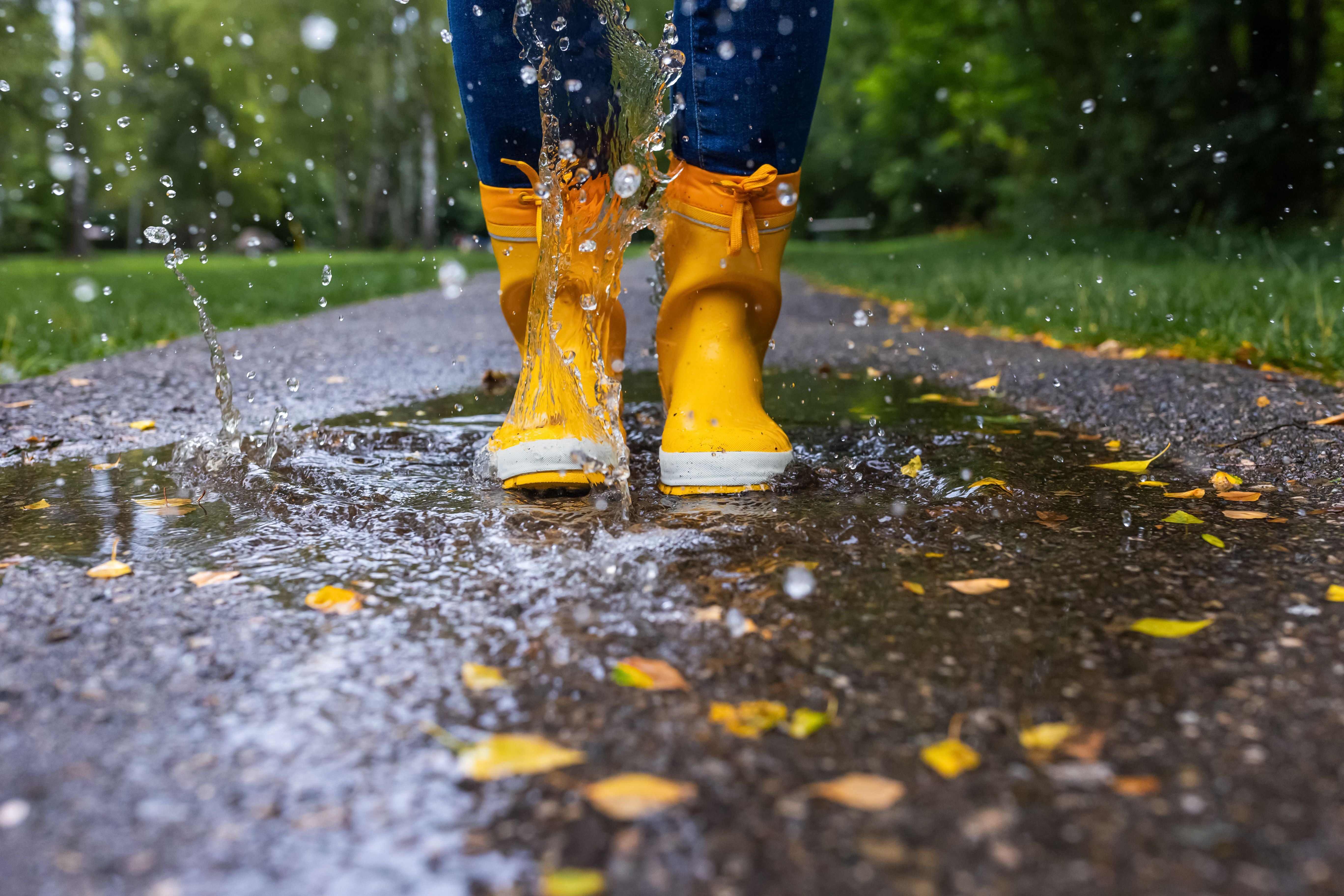 Person mit gelben Gummistiefeln springt in eine Pfütze auf einem Weg im Park.