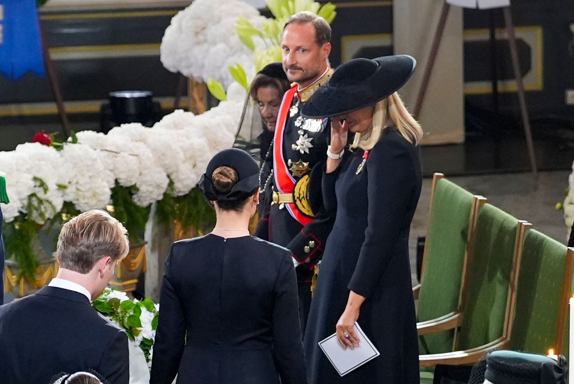 Menschen in schwarzer Trauerkleidung bei einer Trauerfeier mit weißen Blumen und grünen Stühlen.