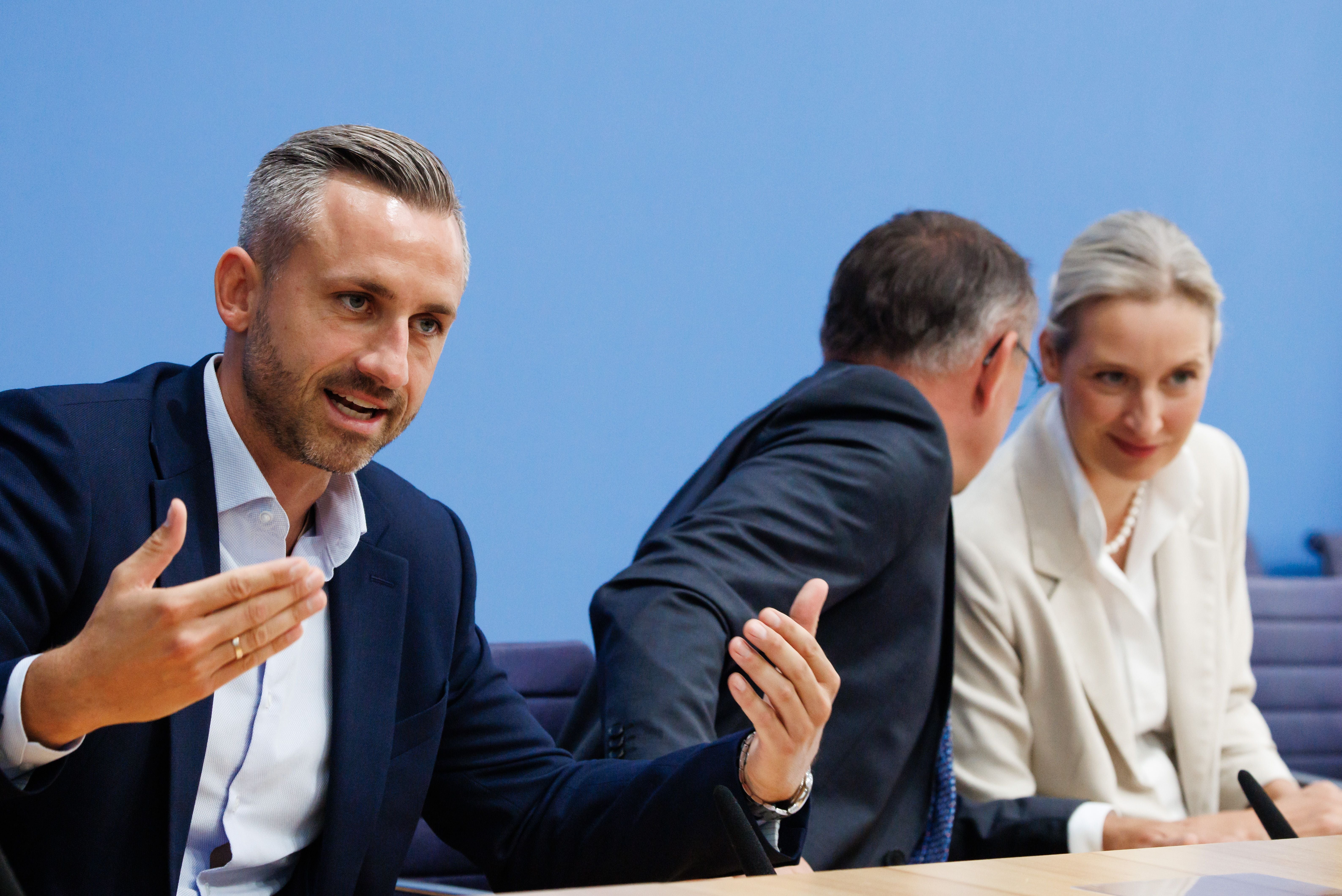 Drei Personen im Anzug sprechen auf einer Pressekonferenz vor blauem Hintergrund.