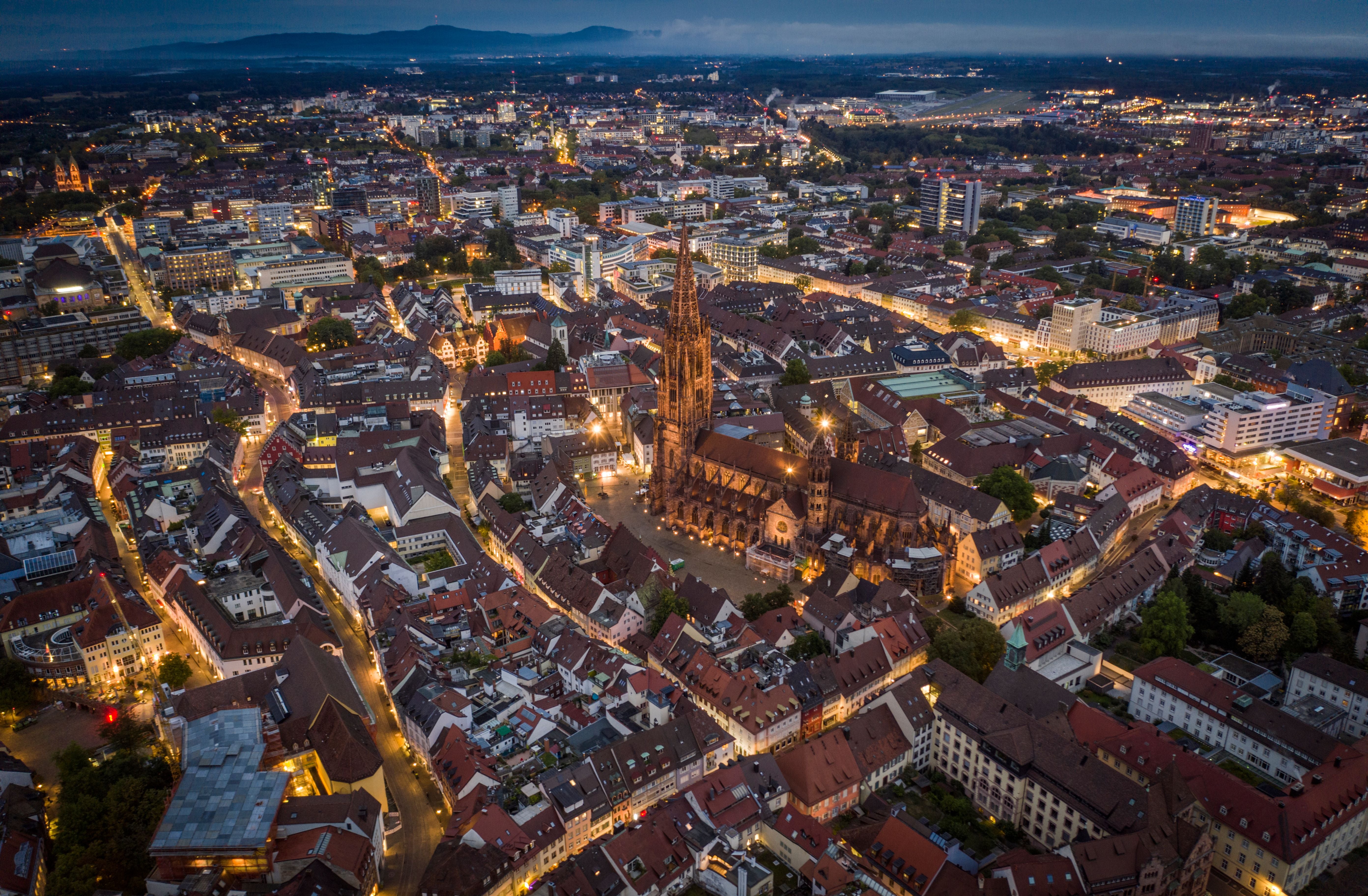 Luftaufnahme von Freiburg im Breisgau mit beleuchtetem Münster und umgebenden Häusern bei Dämmerung.