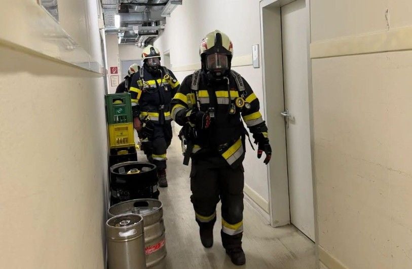 Zwei Feuerwehrleute mit Atemschutzmasken gehen durch einen engen Hotelflur mit Fässern.