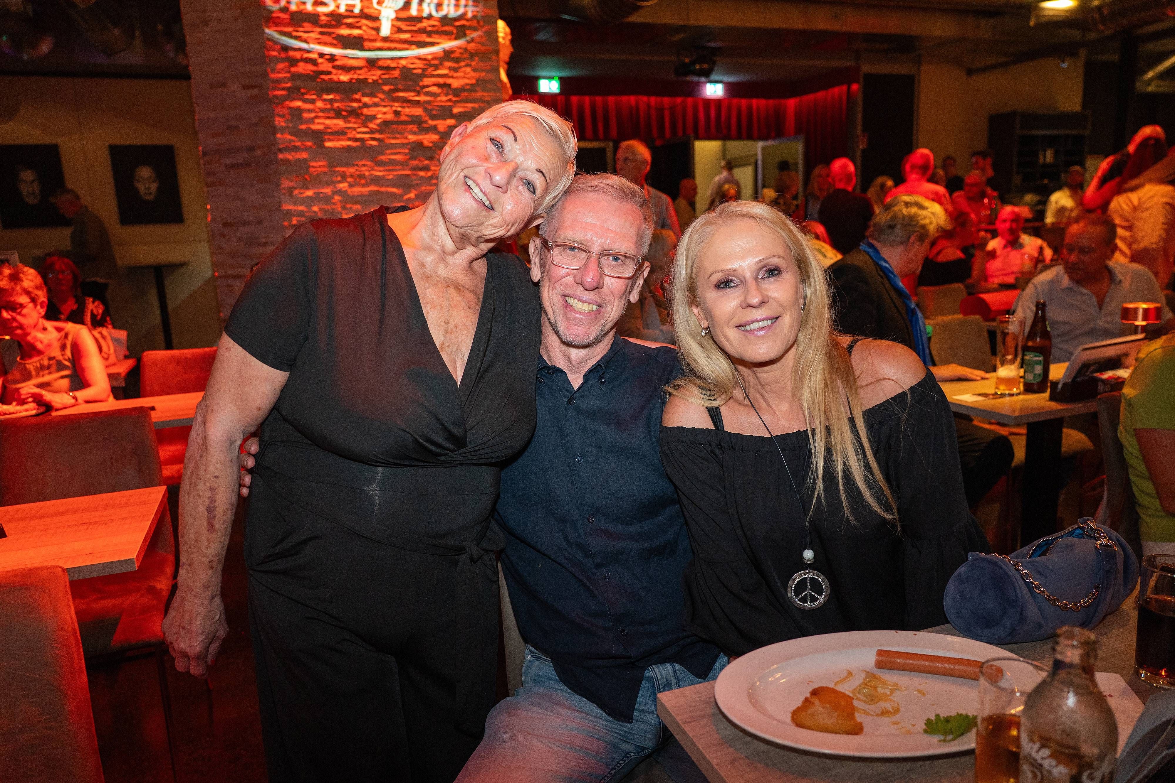 Drei Menschen lächeln in einem vollen Restaurant bei der Premiere von "The Sound of Strobl".