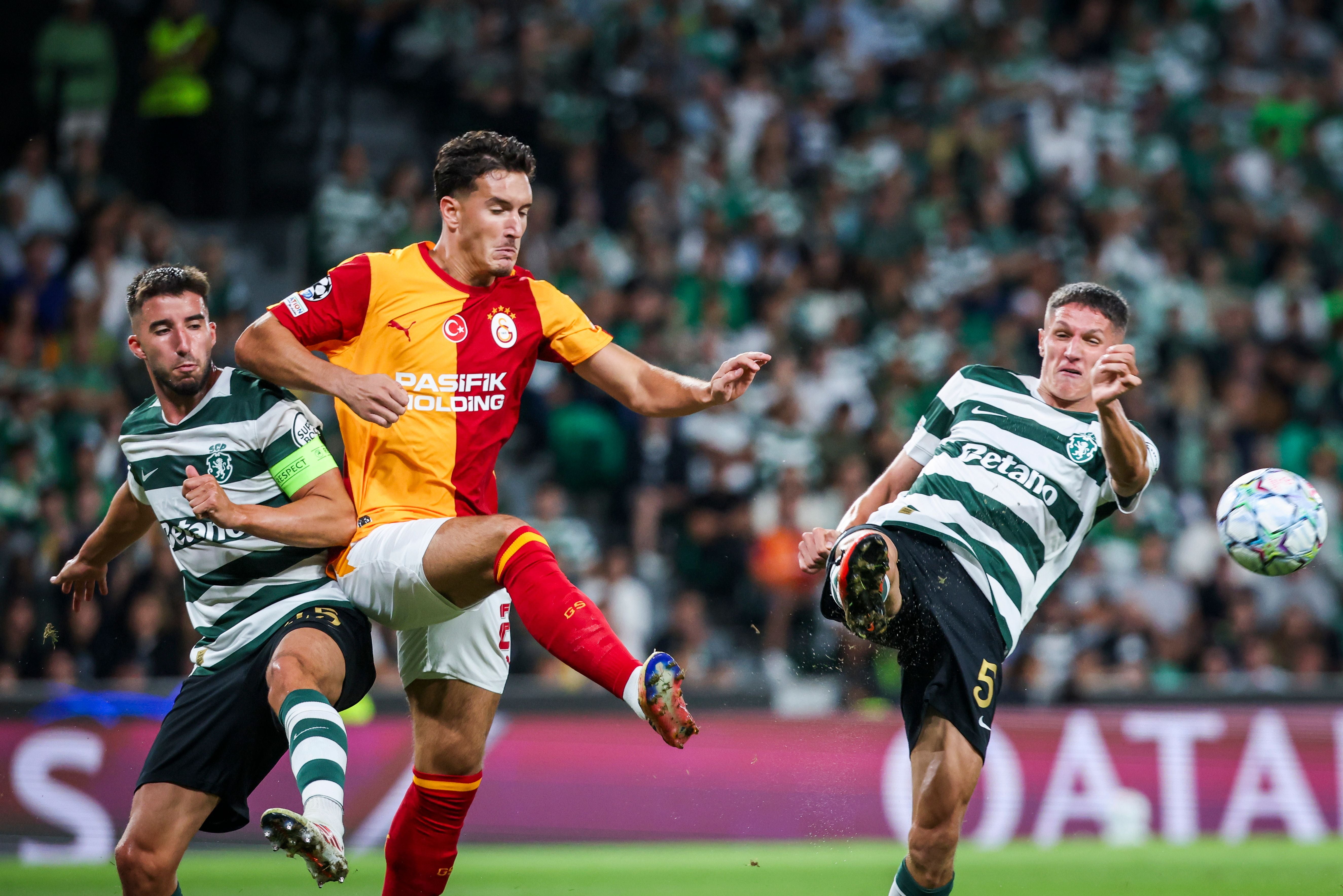 Ein Fußballspieler von Galatasaray Istanbul, in gelb-rotem Trikot, holt zum Schuss aus. Ihm stehen zwei Gegenspieler von Sporting Lissabon, in grün-weiß gestreiften Trikots, im Weg, von denen einer grätscht. Die Szene spielt sich vor einer vollen Fantribüne ab.