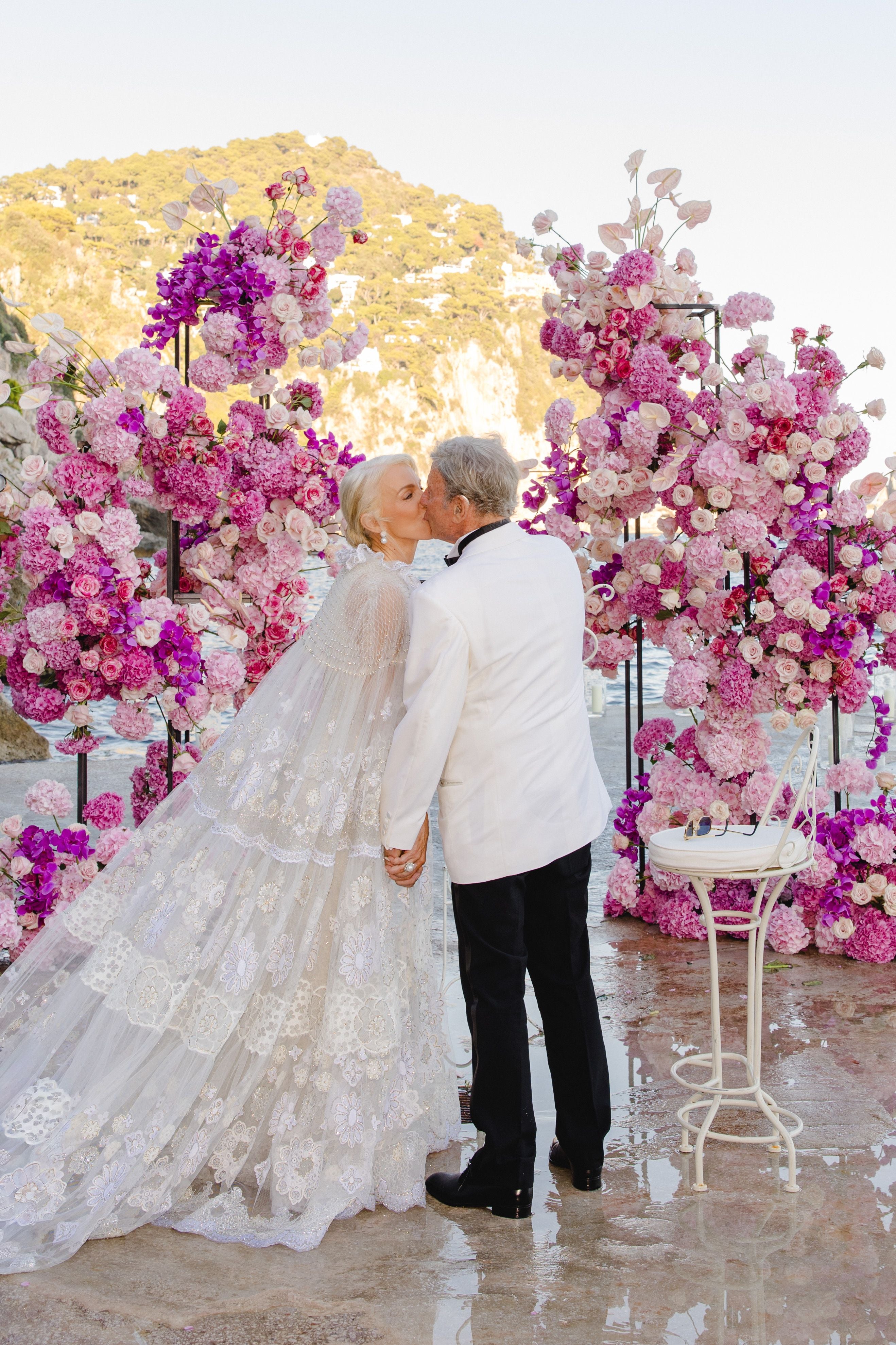 Ein älteres Paar küsst sich vor rosa Blumen bei einer Hochzeit im Freien.