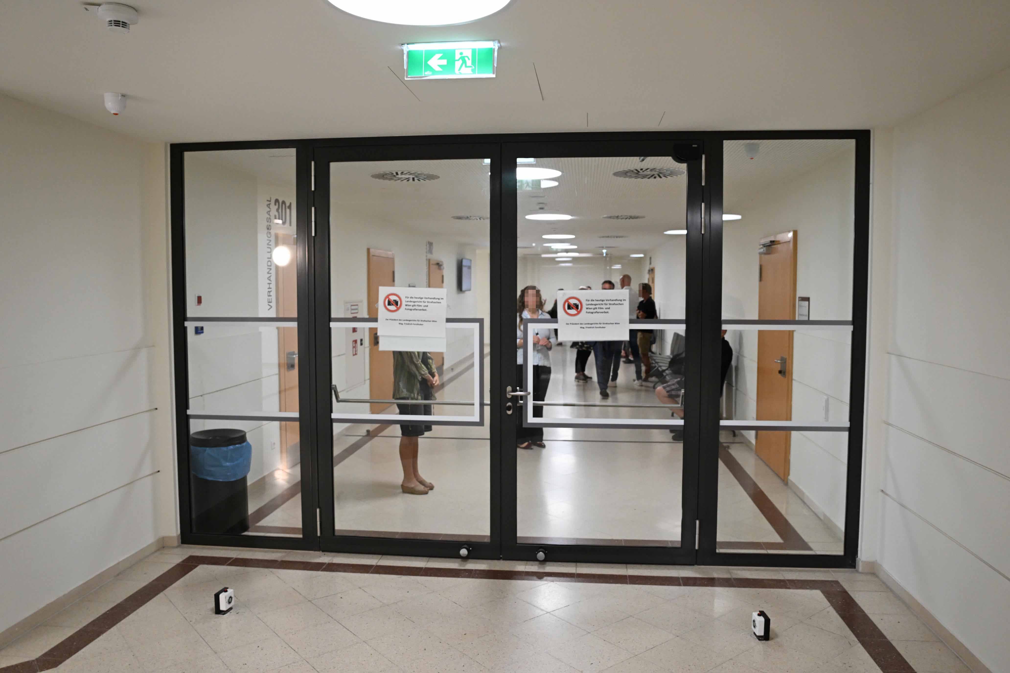 Glasflurtür mit Hinweisschildern, dahinter Personen im Gang eines öffentlichen Gebäudes.