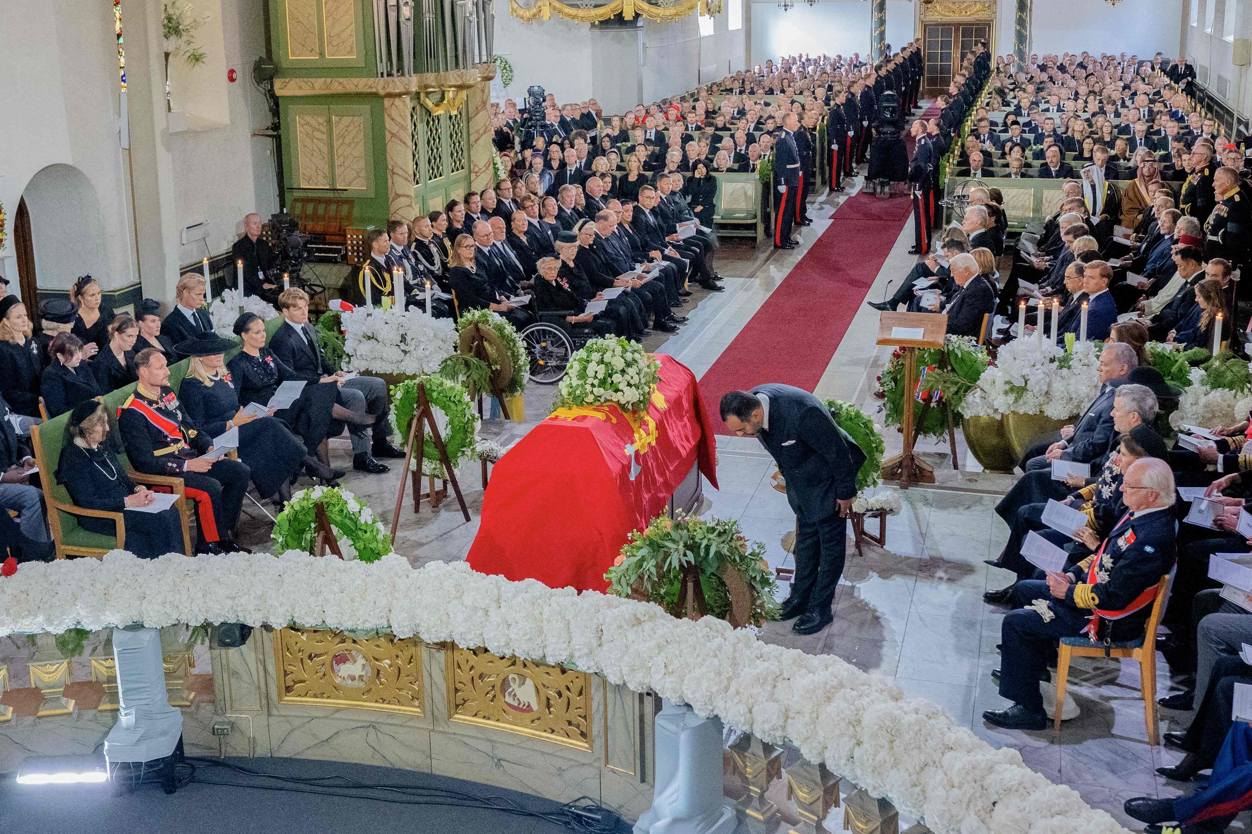 Trauergottesdienst mit rotem Sarg in einer voll besetzten Kirche, Blumen und viele Gäste in dunkler Kleidung.