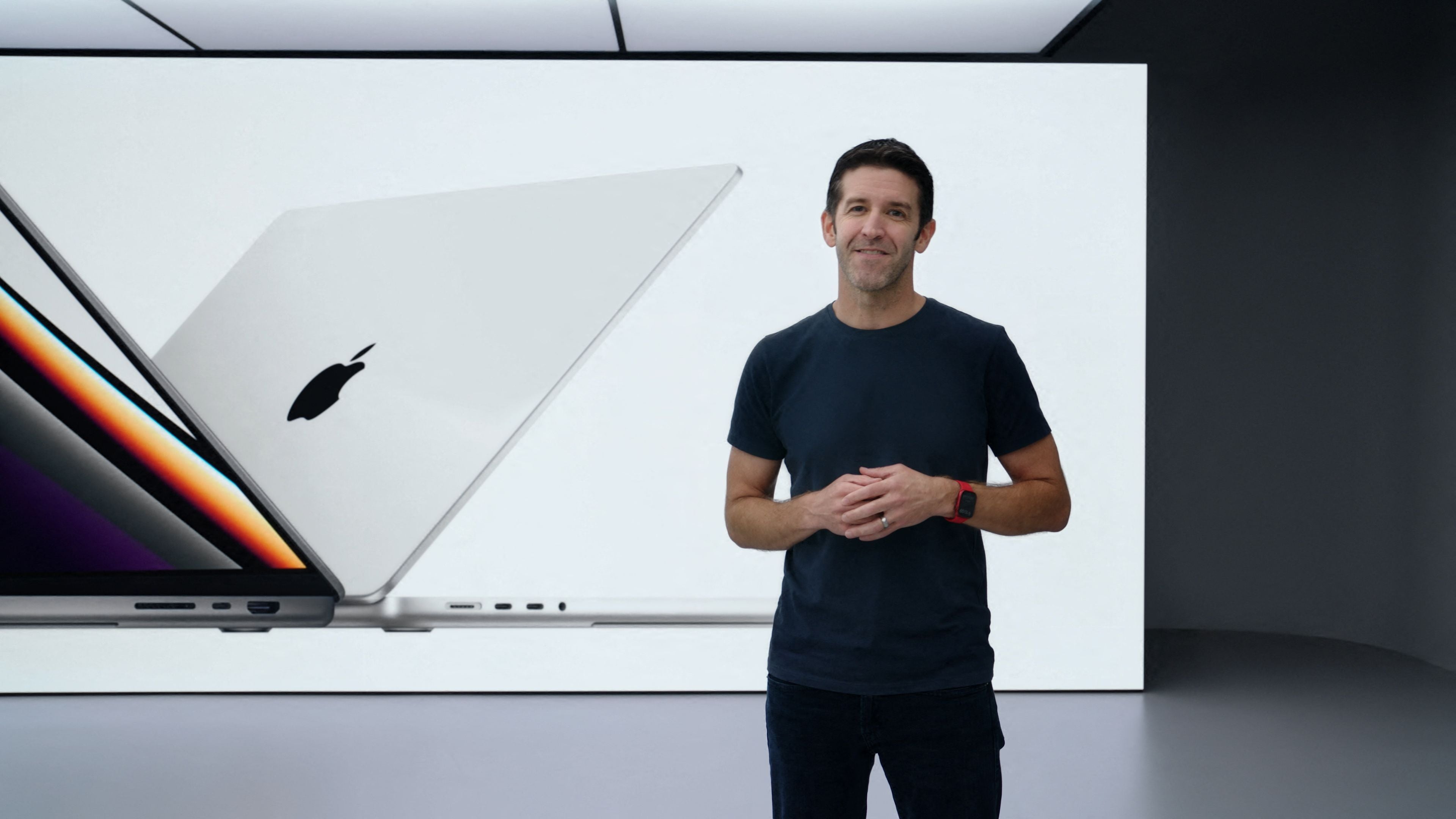 Ein Mann steht vor einer großen Präsentation mit einem MacBook Pro auf dem Bildschirm im Hintergrund.
