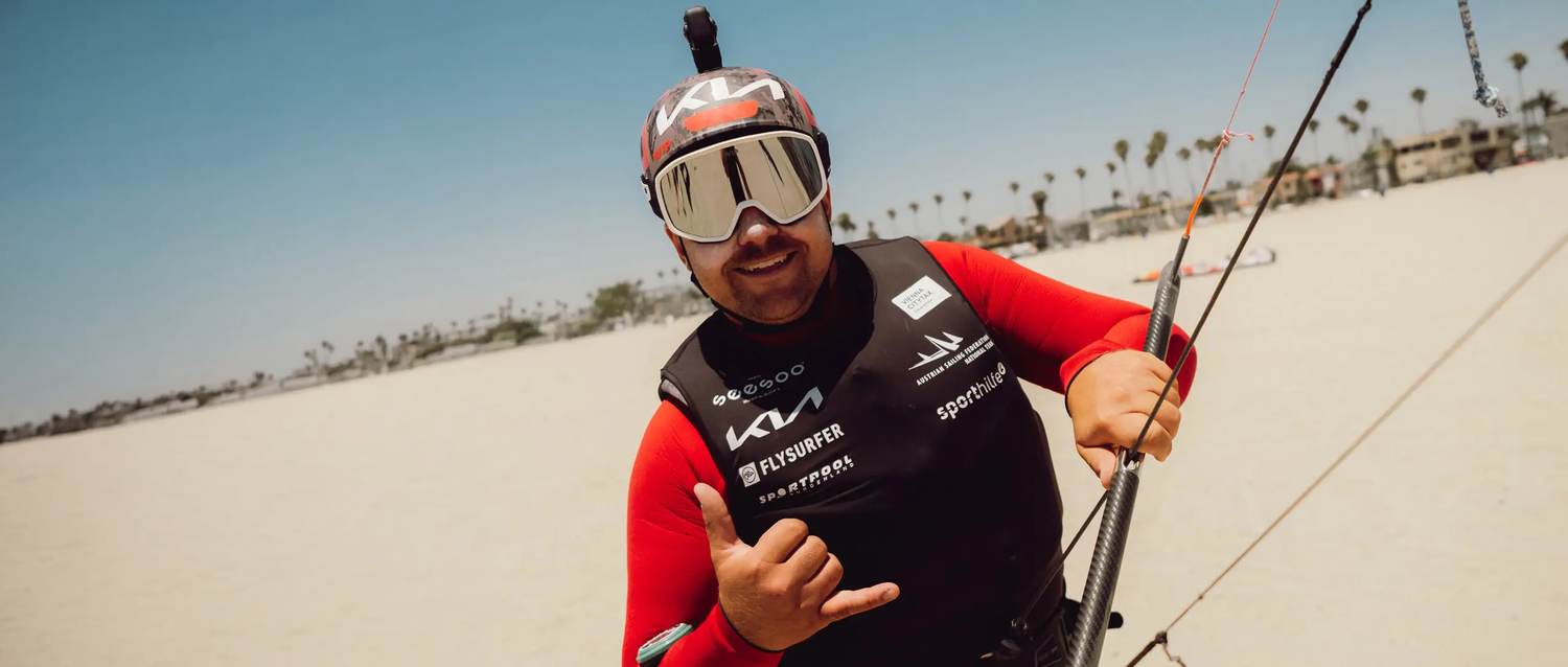 Valentin Bontus im roten Longsleeve, dunkler Schwimmweste und Helm mit verspiegelter Skibrille lächelt und macht am Strand eine Handgeste.