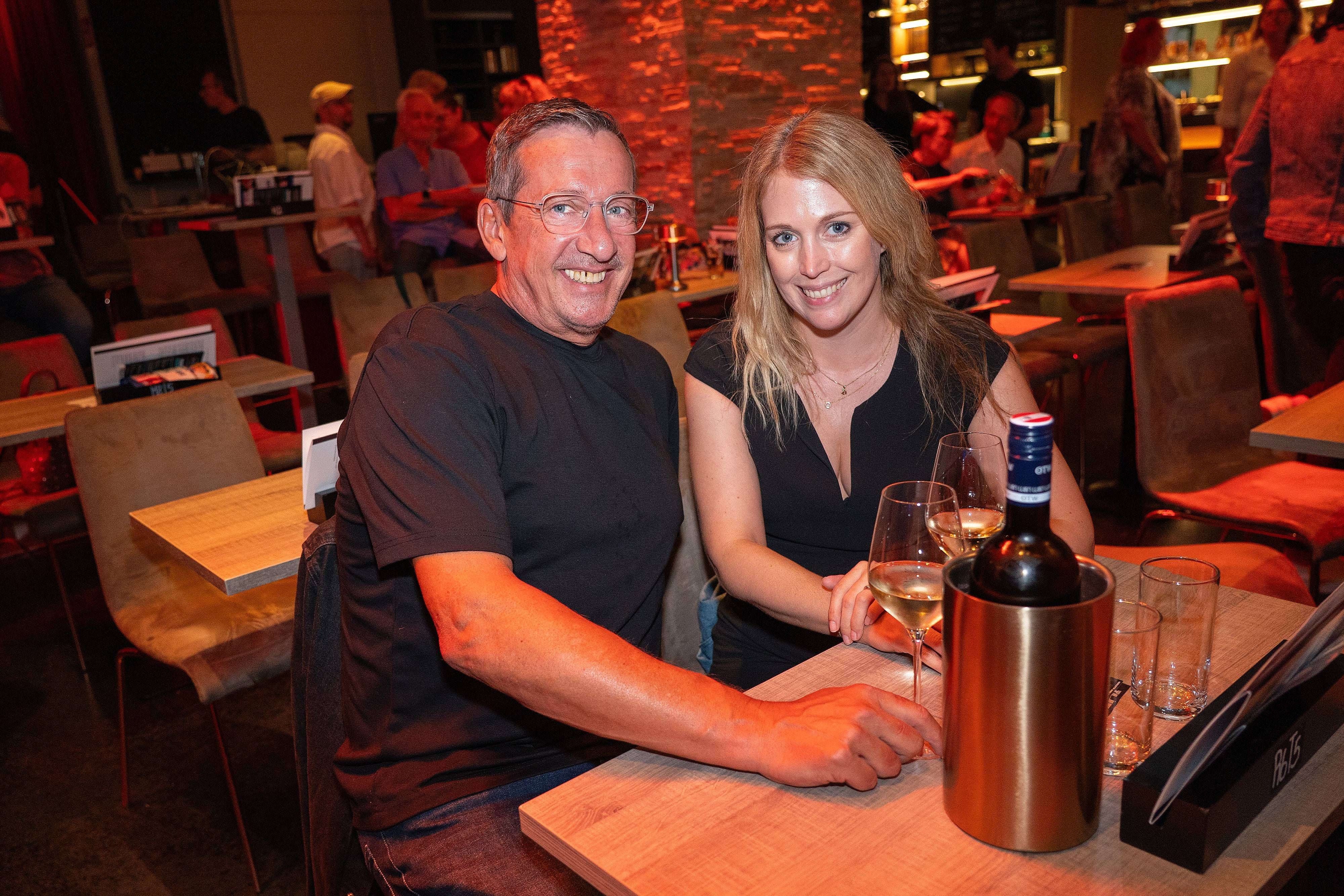 Ein Mann und eine Frau sitzen lachend an einem Restauranttisch mit Wein und Gläsern.