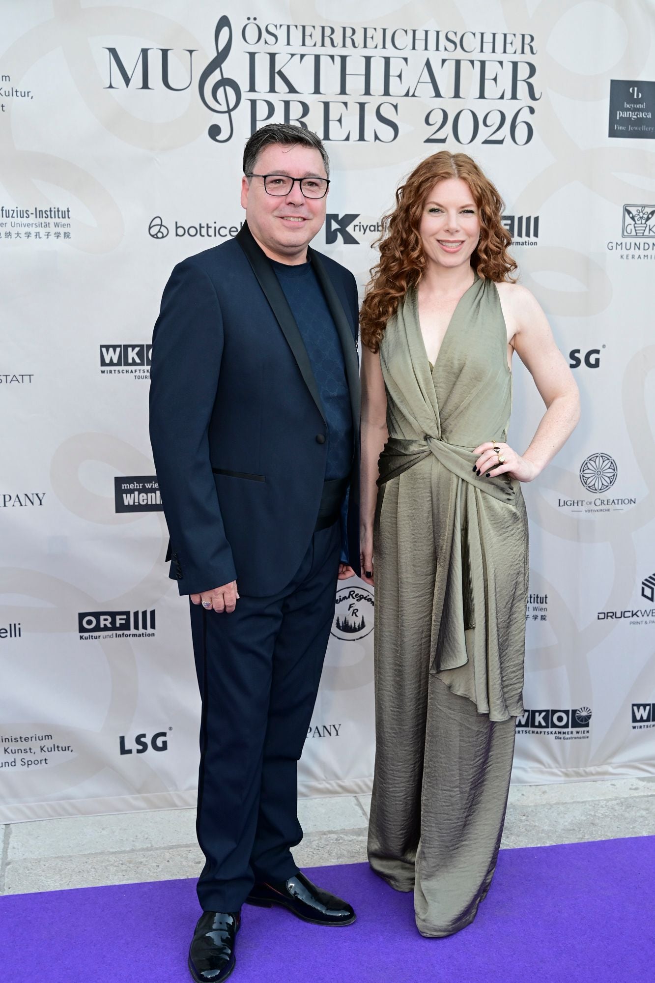 Ein Mann im Anzug und eine Frau im eleganten Kleid posieren auf dem lila Teppich beim Musiktheaterpreis.