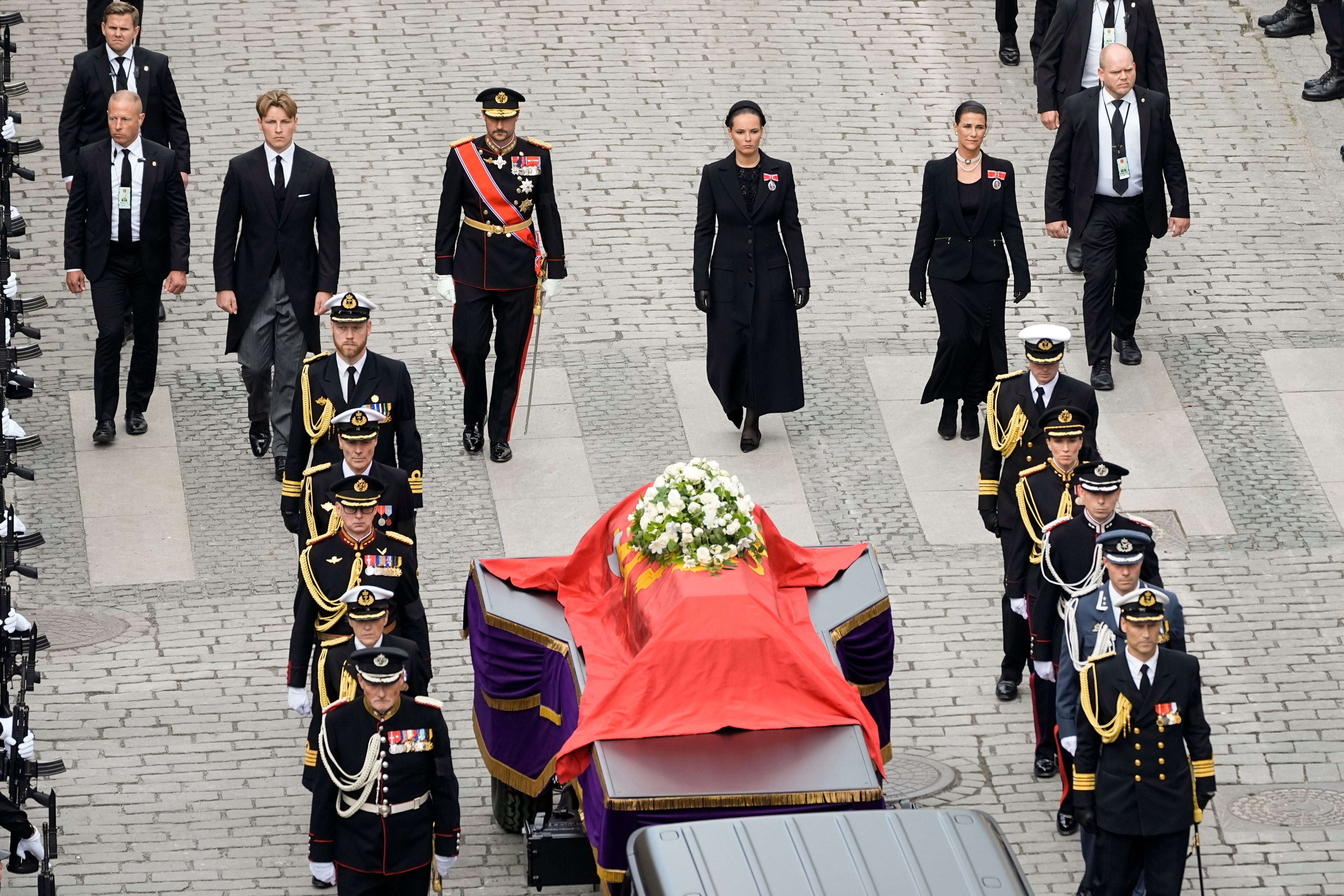 Ein Trauerzug mit Soldaten und trauernden Royals begleitet einen Sarg, bedeckt mit rotem Tuch und Blumen.