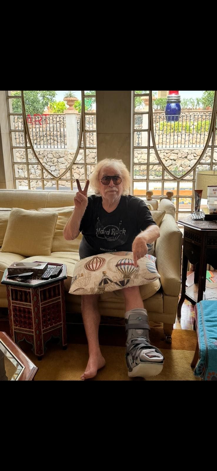 Älterer Mann mit Gipsverband am Fuß sitzt auf Sofa und zeigt das Peace-Zeichen.