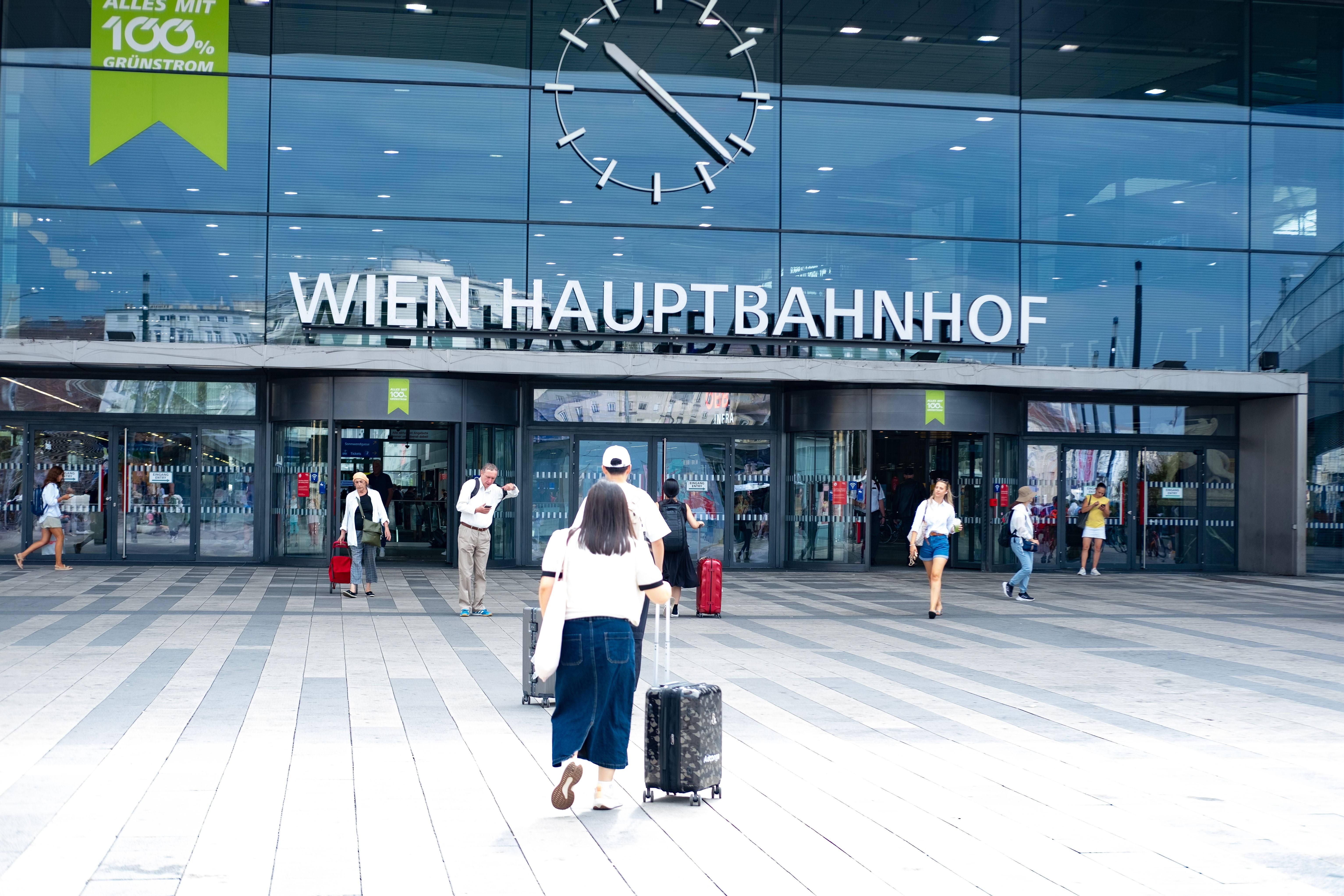 Menschen mit Koffern gehen zum Eingang des Wiener Hauptbahnhofs mit moderner Glasfassade.