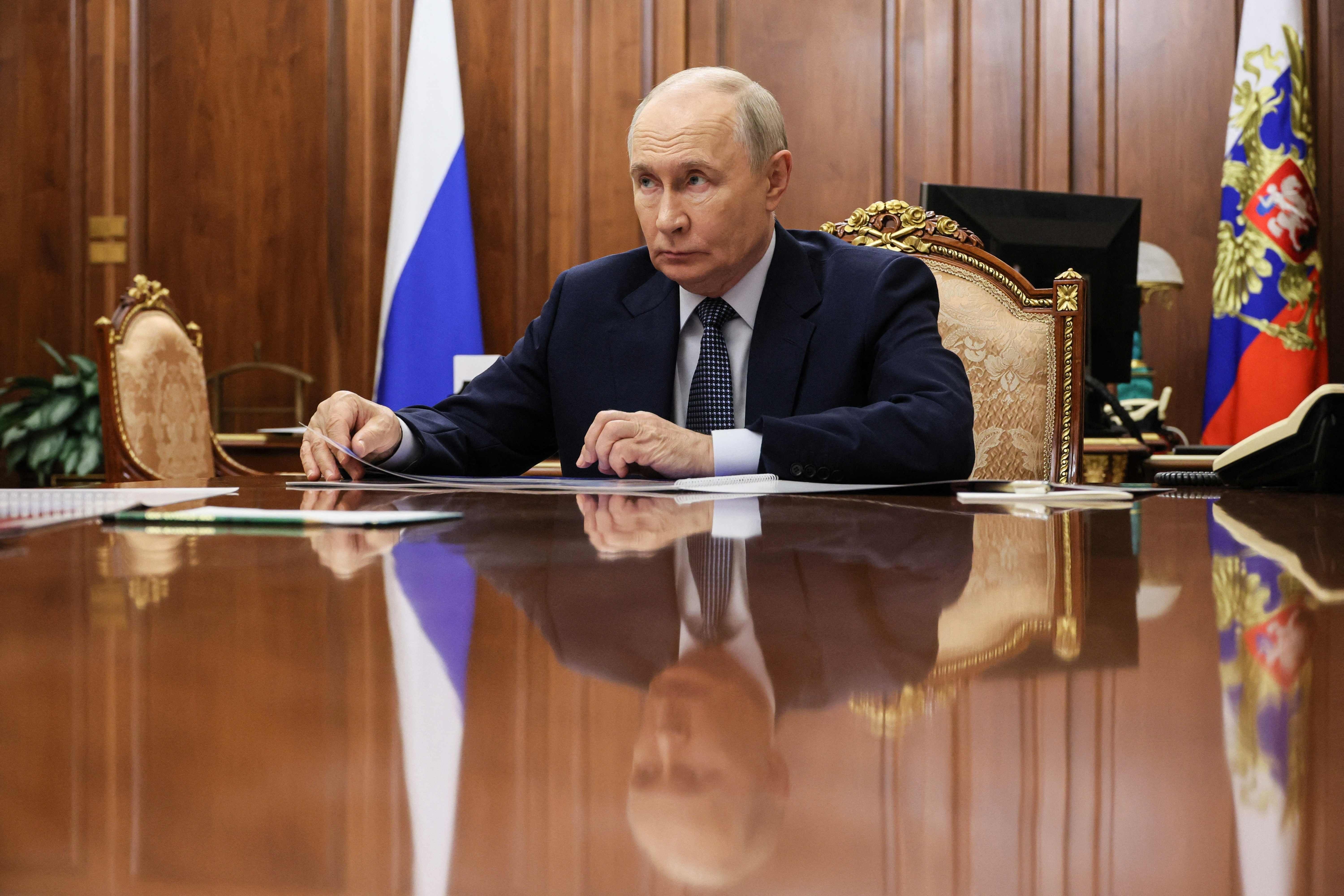 Ein Mann im Anzug sitzt an einem großen Tisch mit russischen Flaggen im Hintergrund.