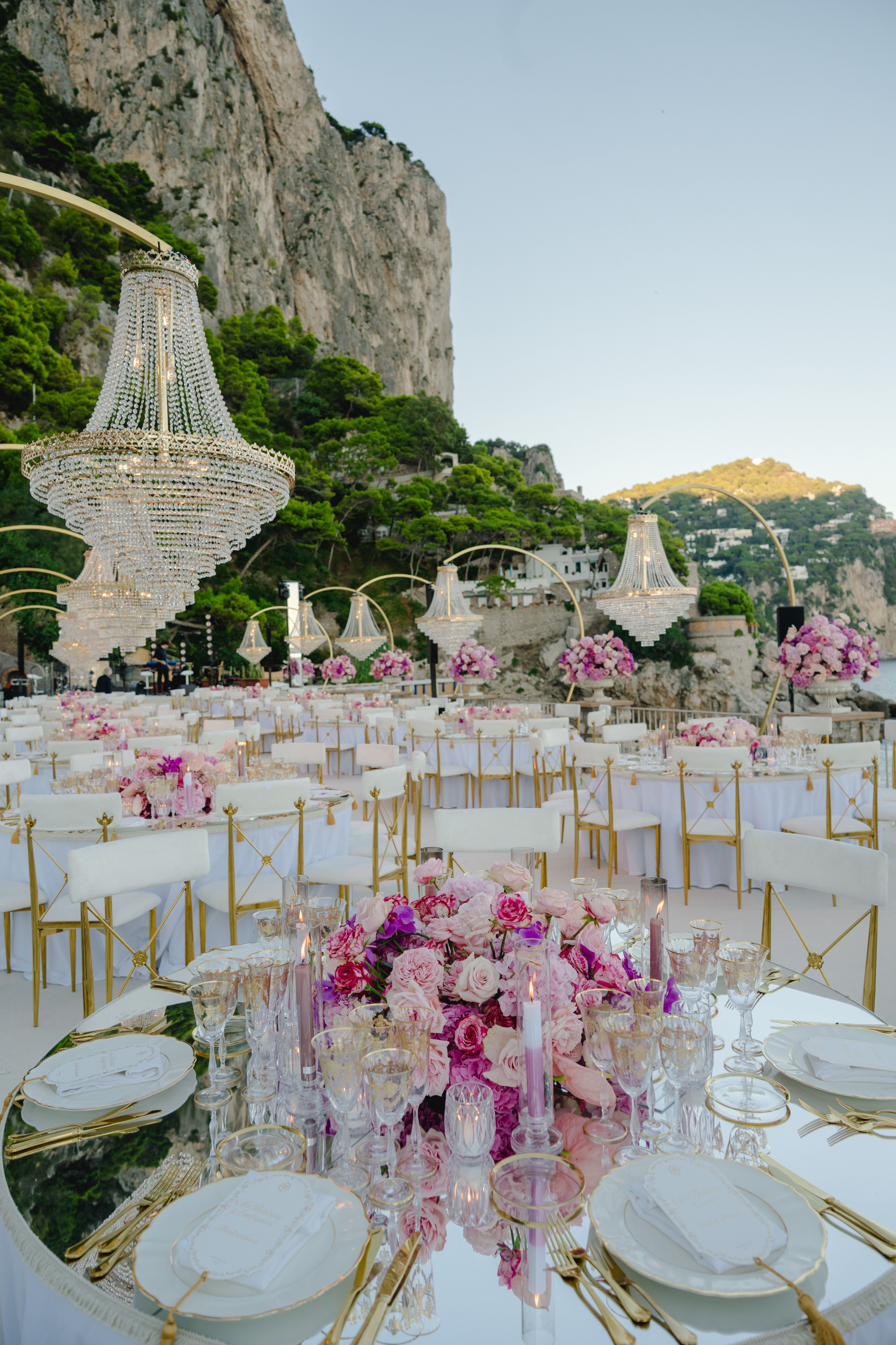 Elegant gedeckte Tische mit rosa Blumen, Kronleuchtern und Felsen im Hintergrund im Freien.