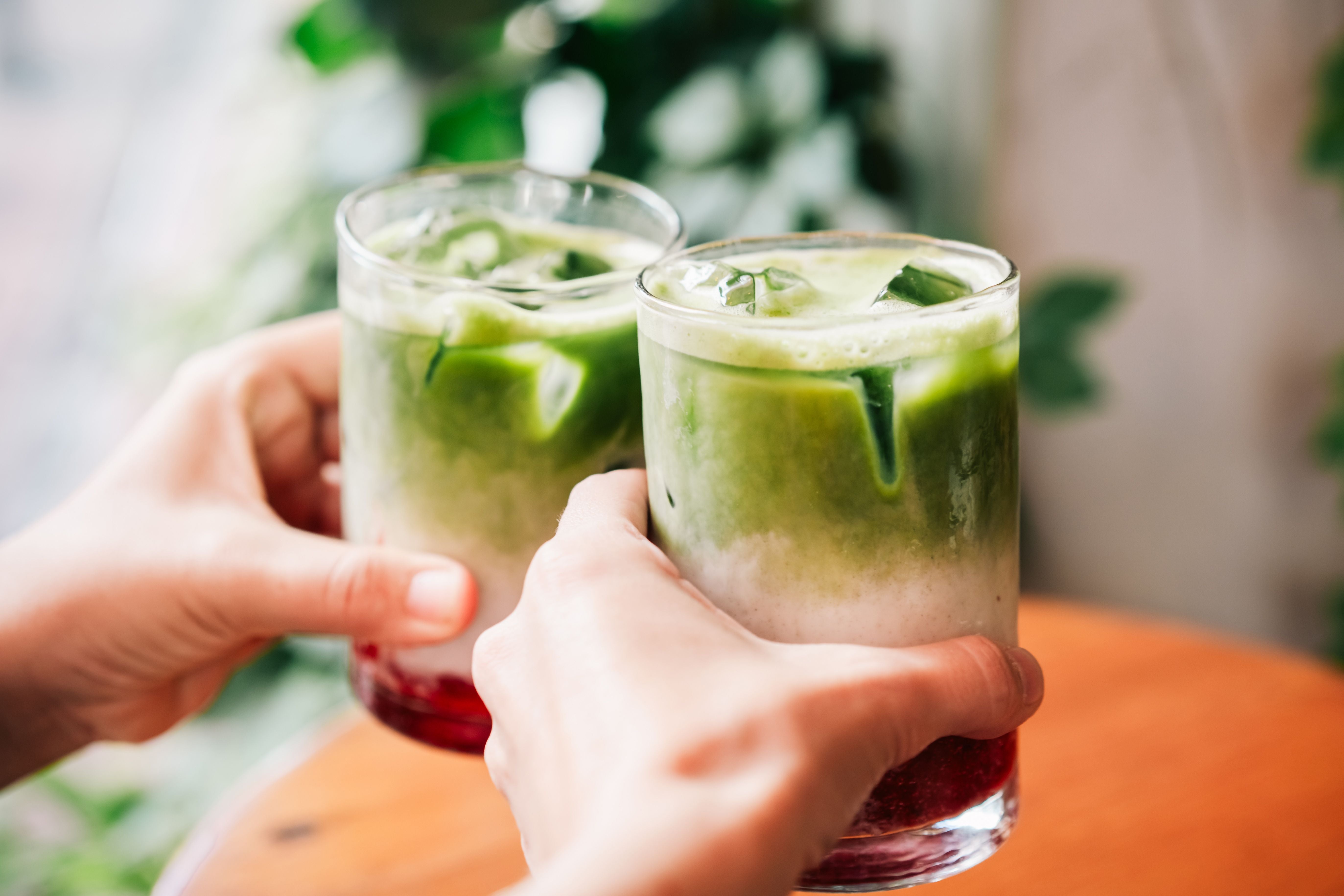 Zwei Hände stoßen mit Gläsern voller Matcha-Latte mit Eis und Beeren-Sirup an.