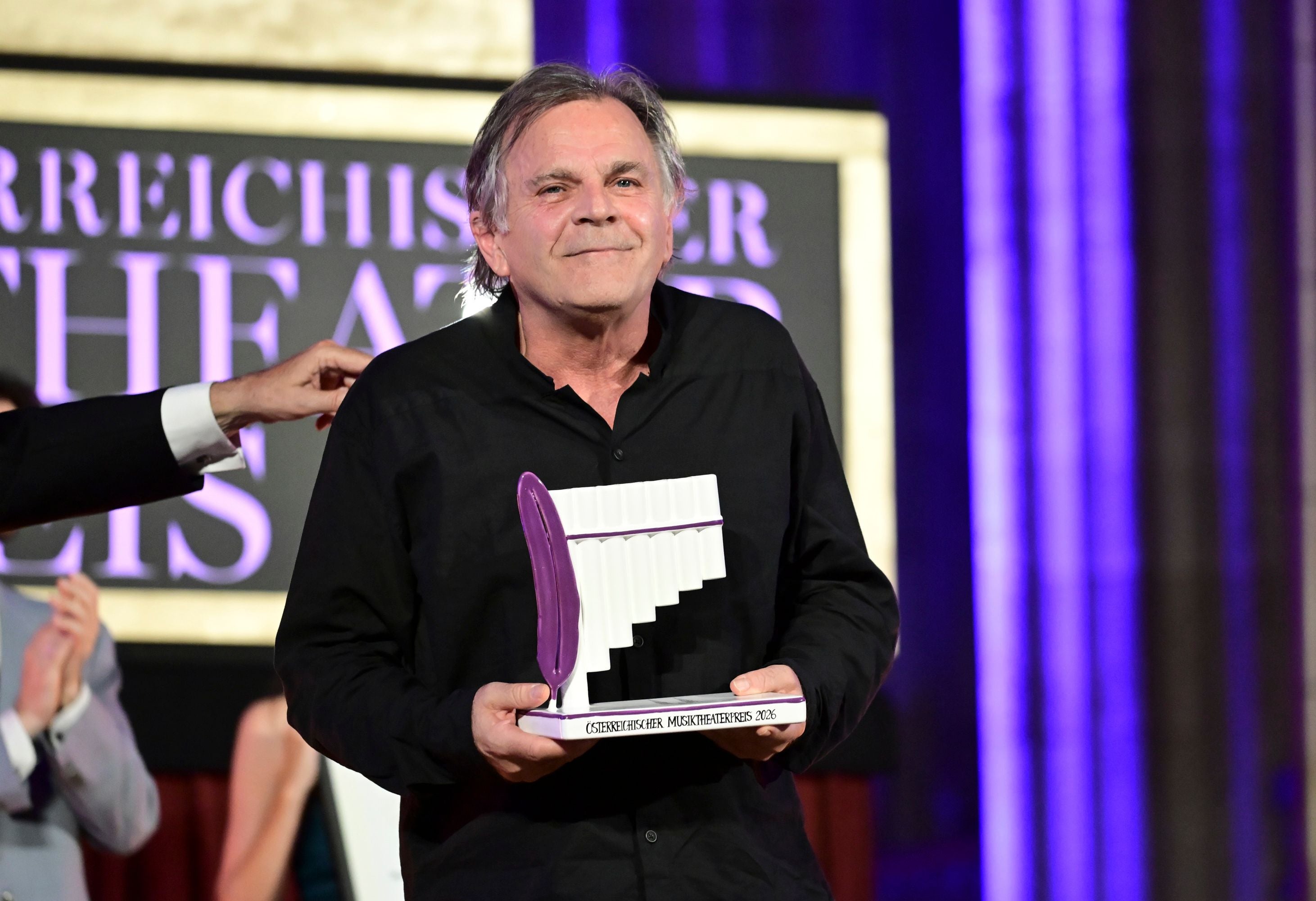 Ein Mann hält den Österreichischen Musiktheaterpreis vor einem festlichen Hintergrund.