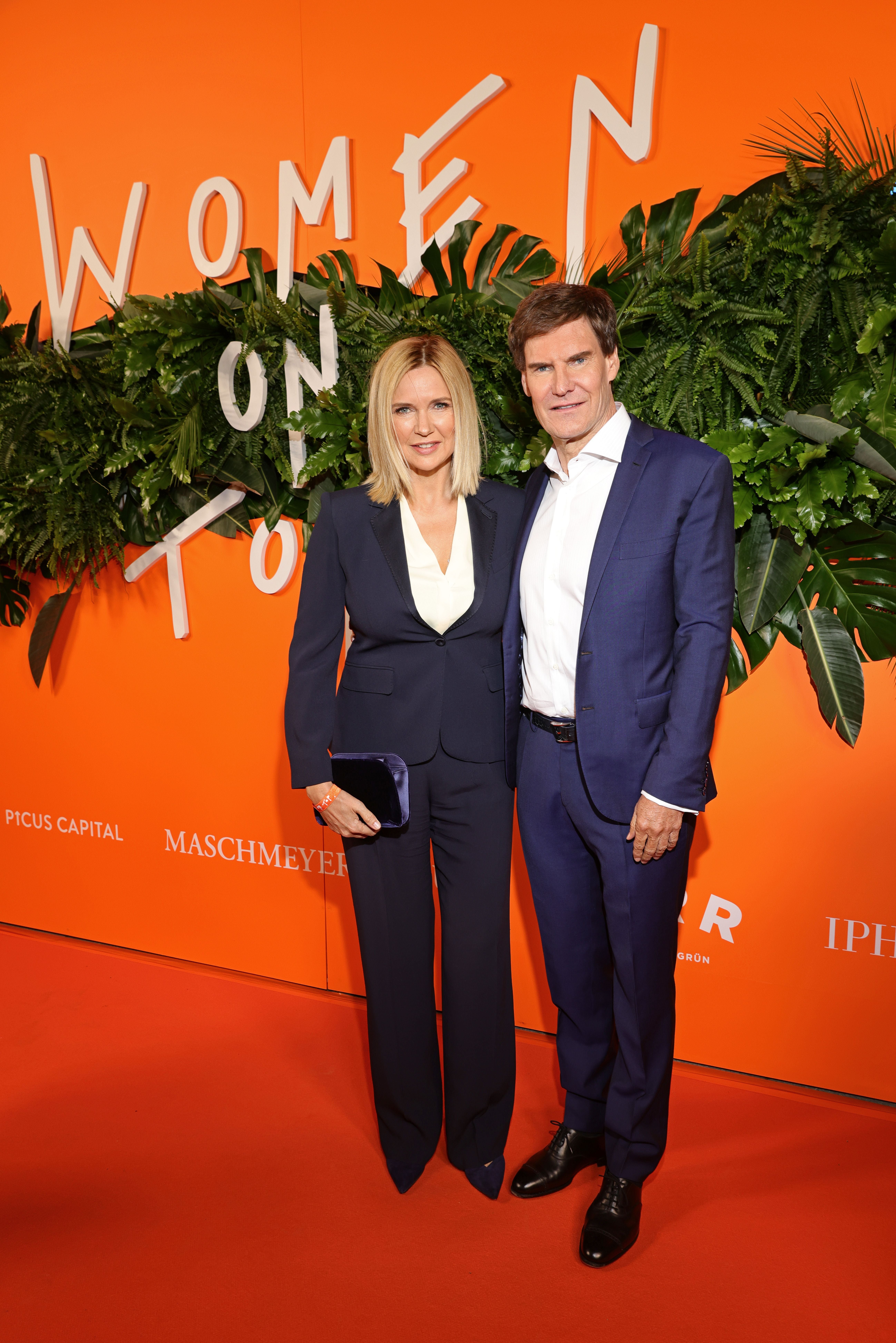 Ein Paar in eleganter Kleidung posiert vor einer orangen "Women On Top"-Wand mit grünen Pflanzen.