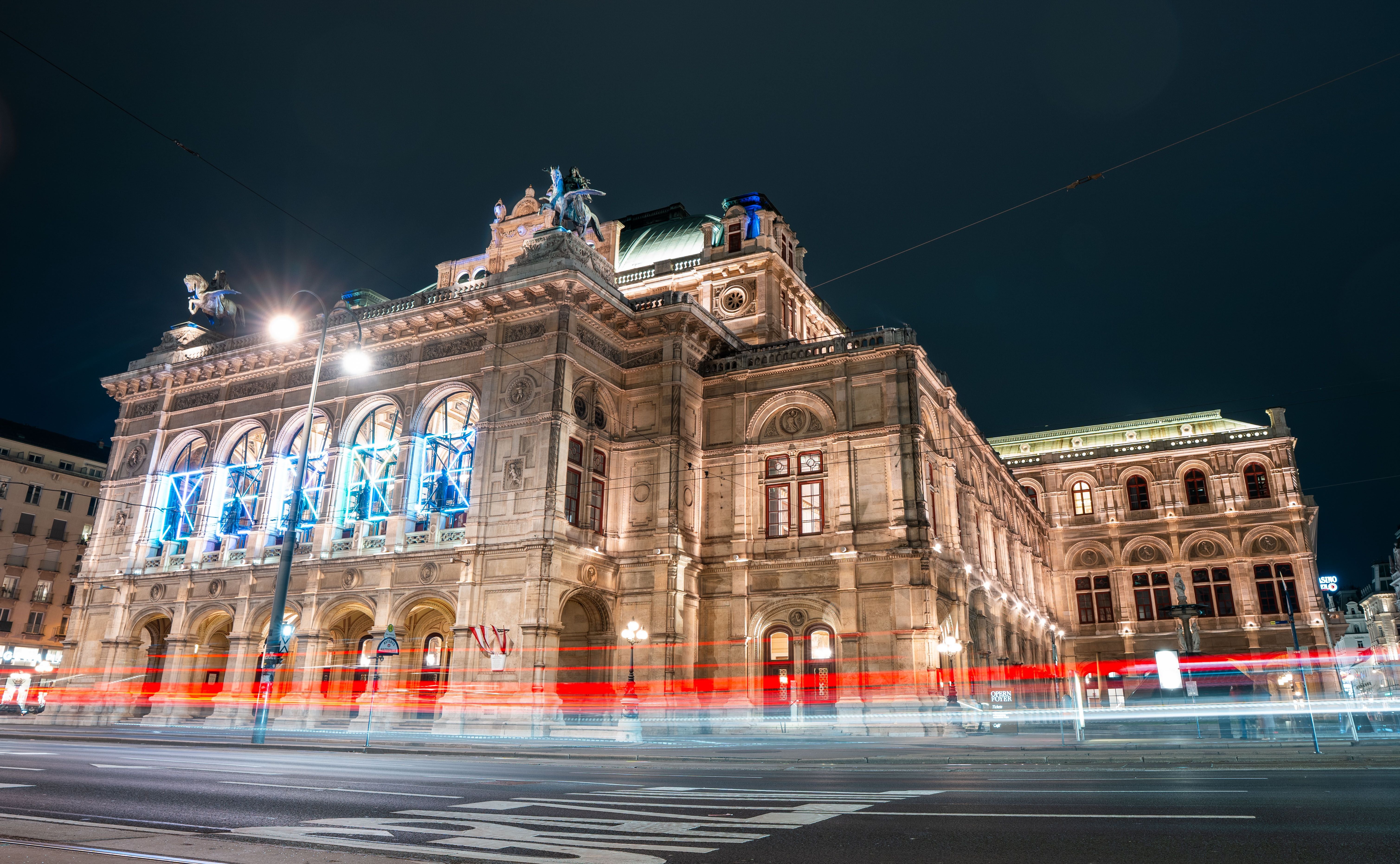 Die Wiener Staatsoper bei Nacht mit beleuchteter Fassade und Lichtspuren vorbeifahrender Autos.