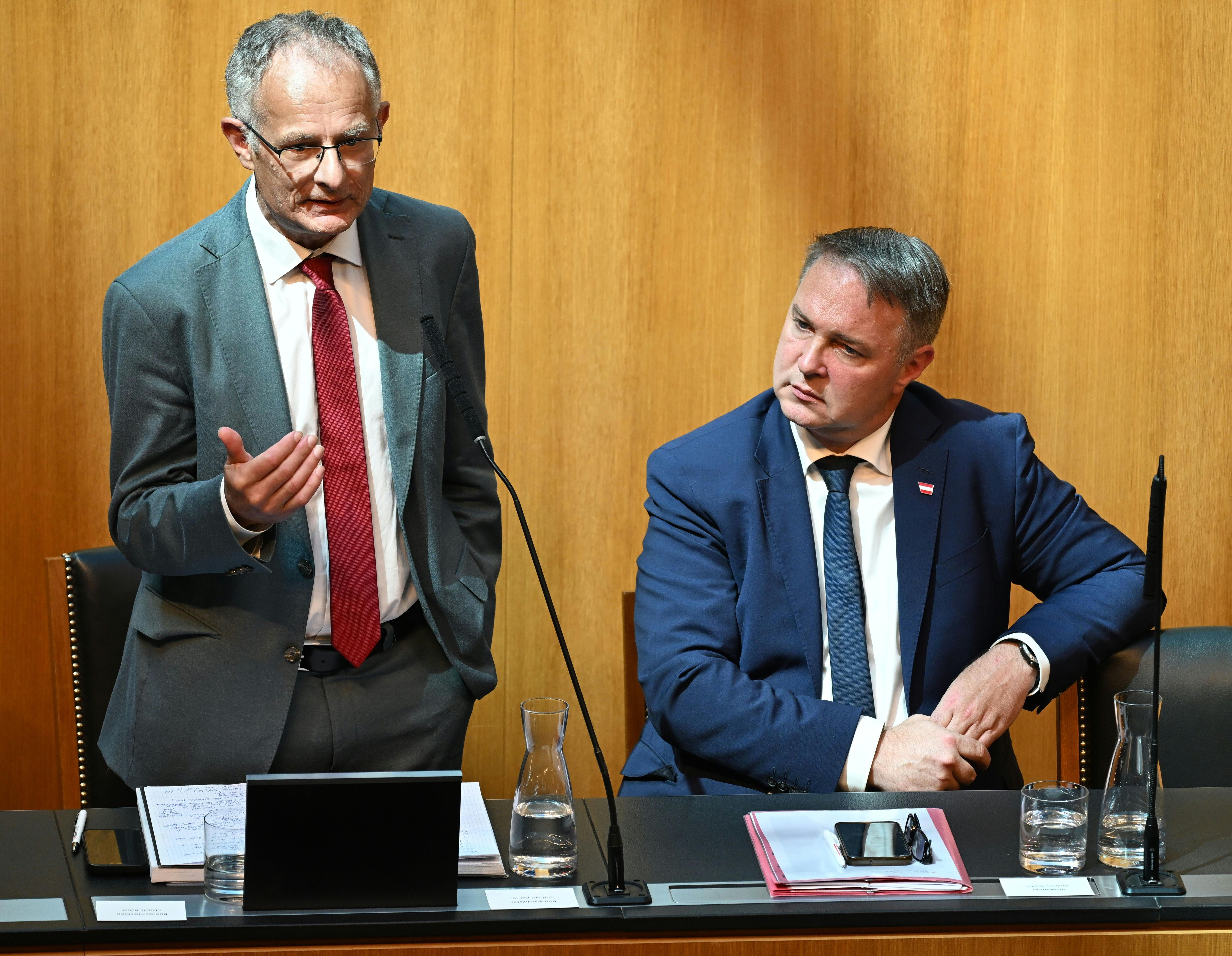 Zwei Männer im Anzug sprechen und sitzen bei einer Parlamentssitzung an einem Tisch.
