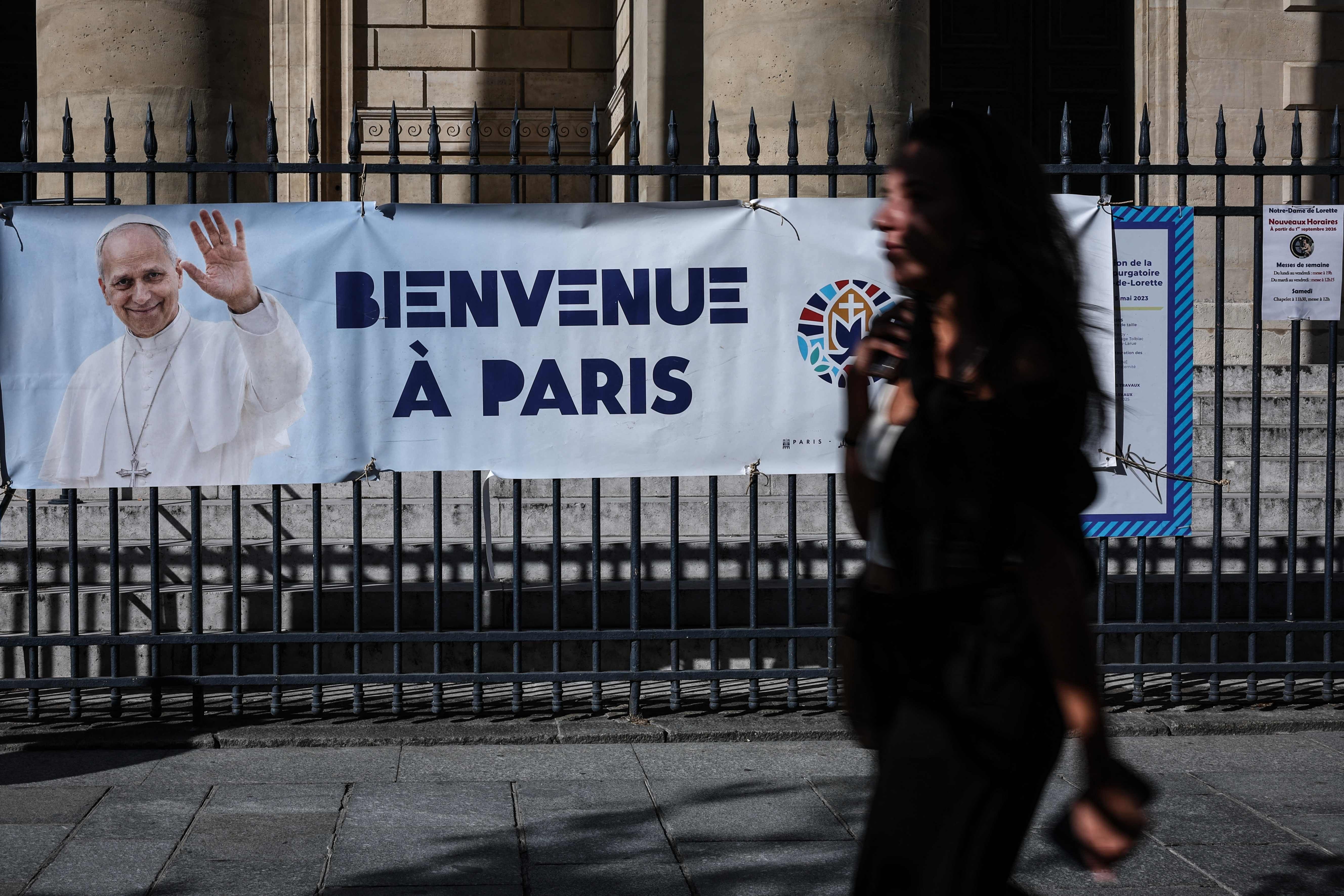 Ein Banner mit Papstbild und "Bienvenue à Paris" hängt vor einer Kirche; eine Person läuft vorbei.