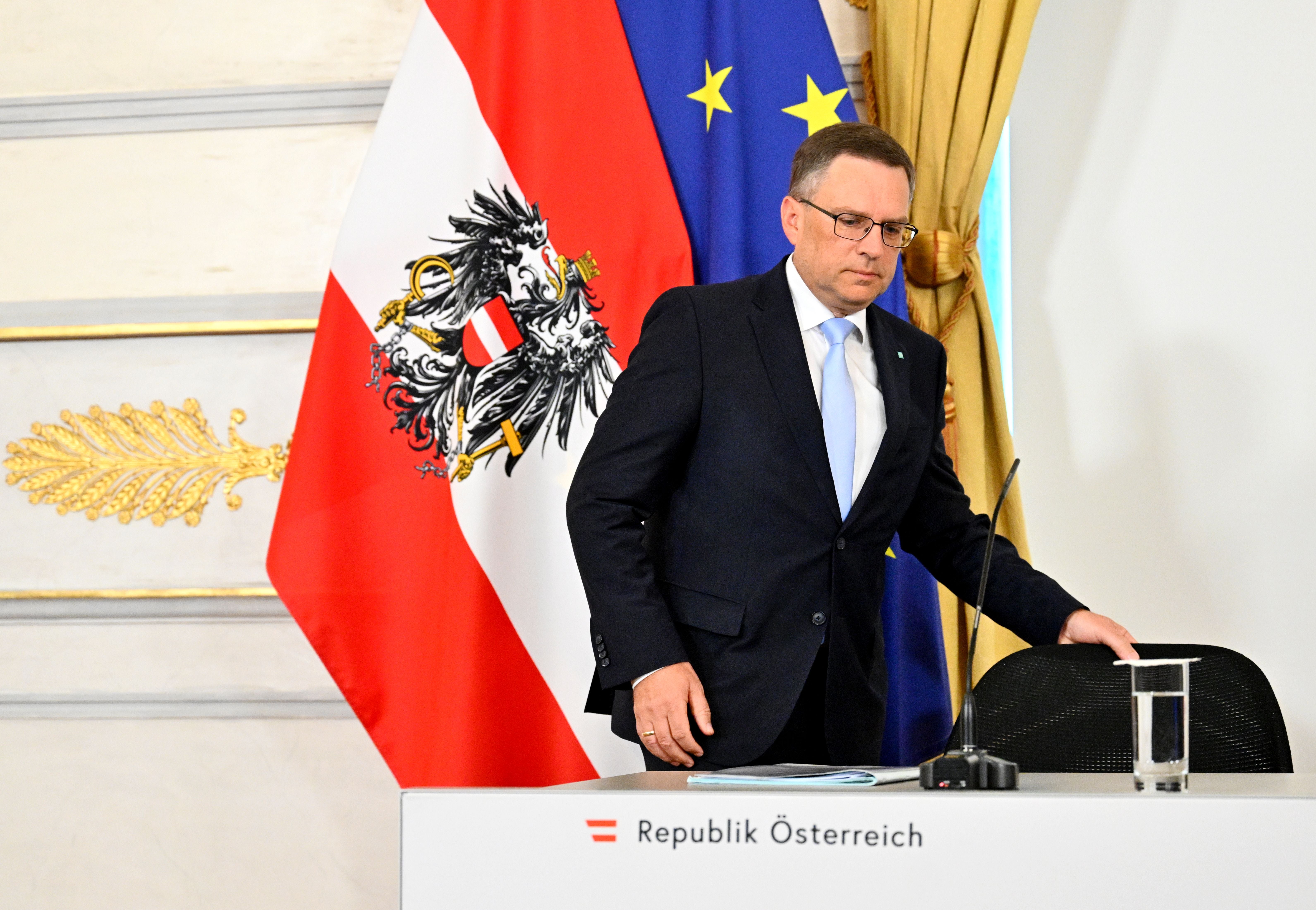 Ein Mann im Anzug steht an einem Tisch mit "Republik Österreich" vor österreichischer Flagge.