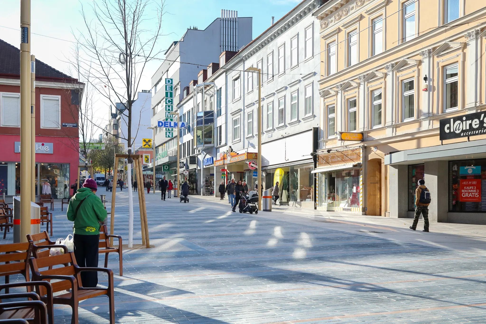 Menschen spazieren auf einer sonnigen Einkaufsstraße mit verschiedenen Geschäften und Bänken.
