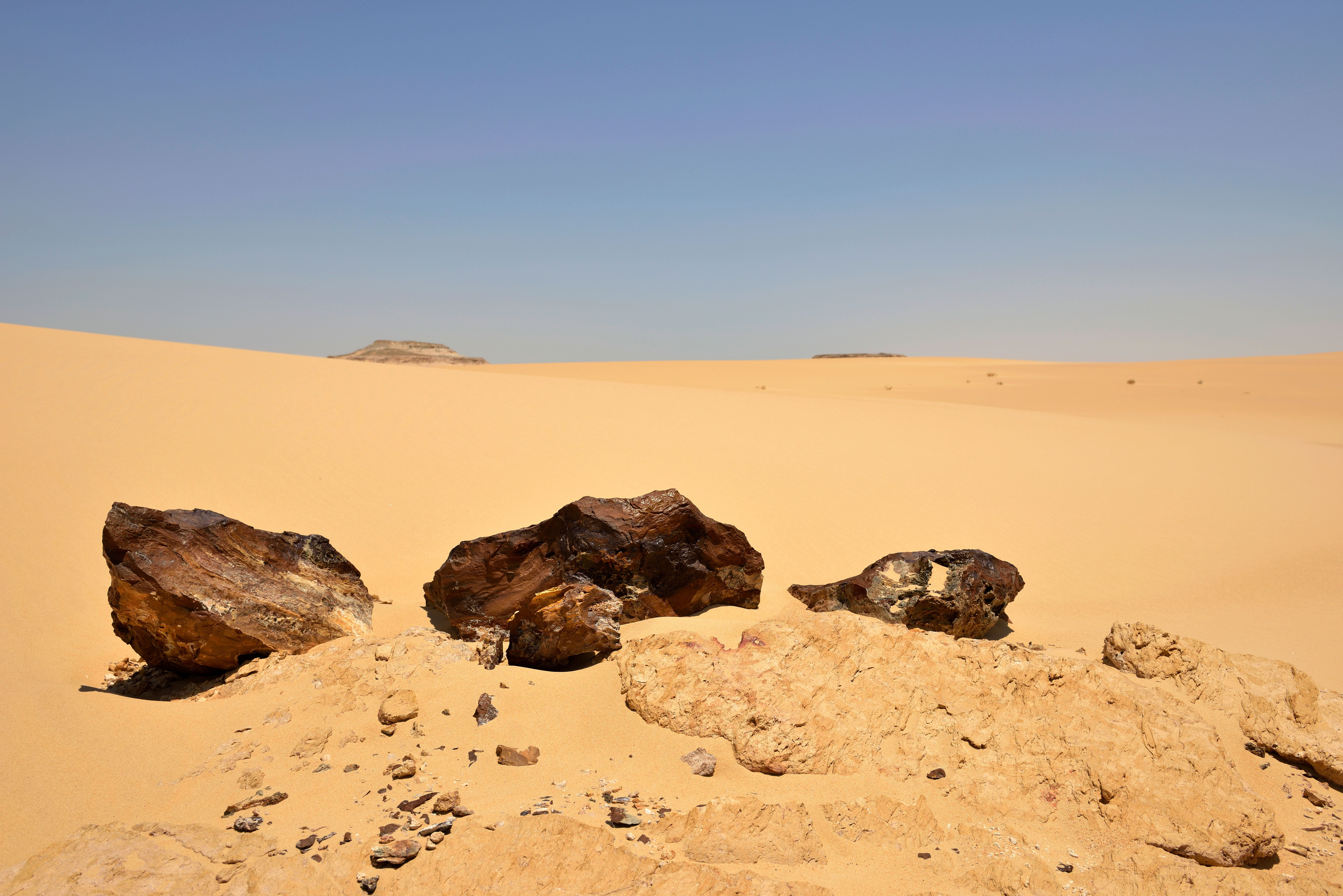 Dunkle Meteoritenstücke liegen auf hellem Sand in der Sahara-Wüste unter klarem Himmel.