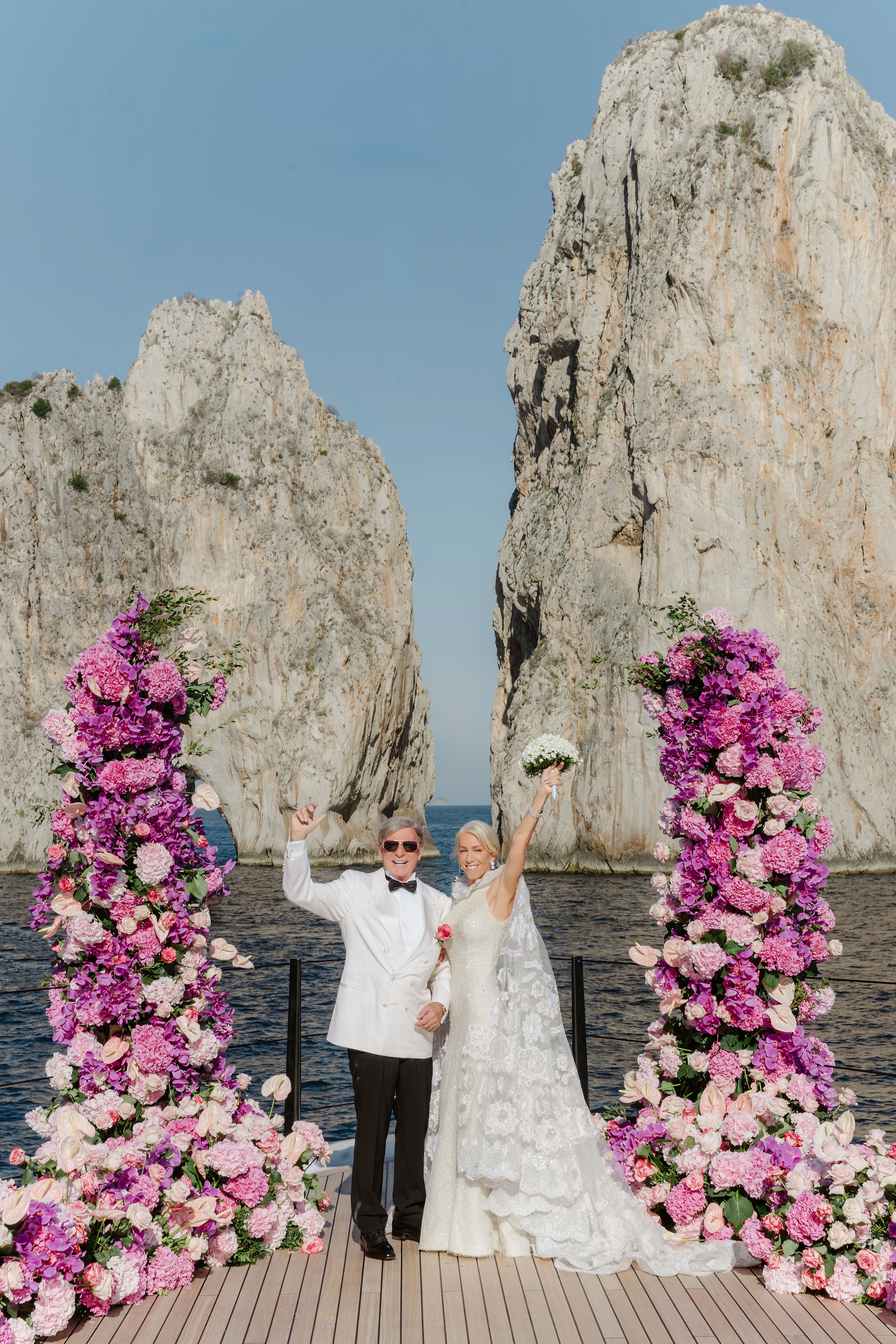 Brautpaar steht zwischen Blumenbögen auf einem Steg vor Felsen am Meer und jubelt.