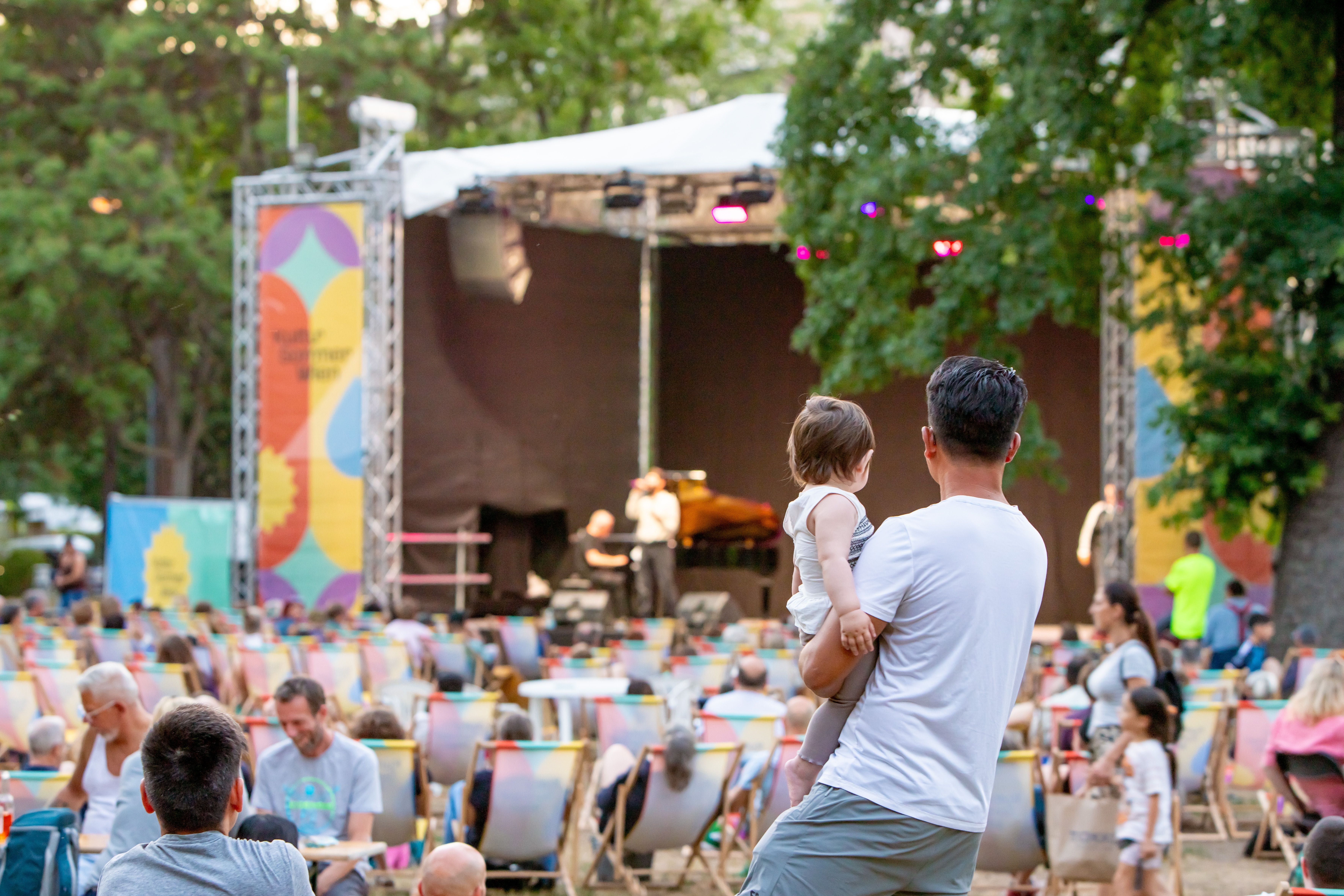 Mehrere Menschen sitzen im Park vor einer Open-Air-Bühne mit Livemusik, Bäume im Hintergrund.