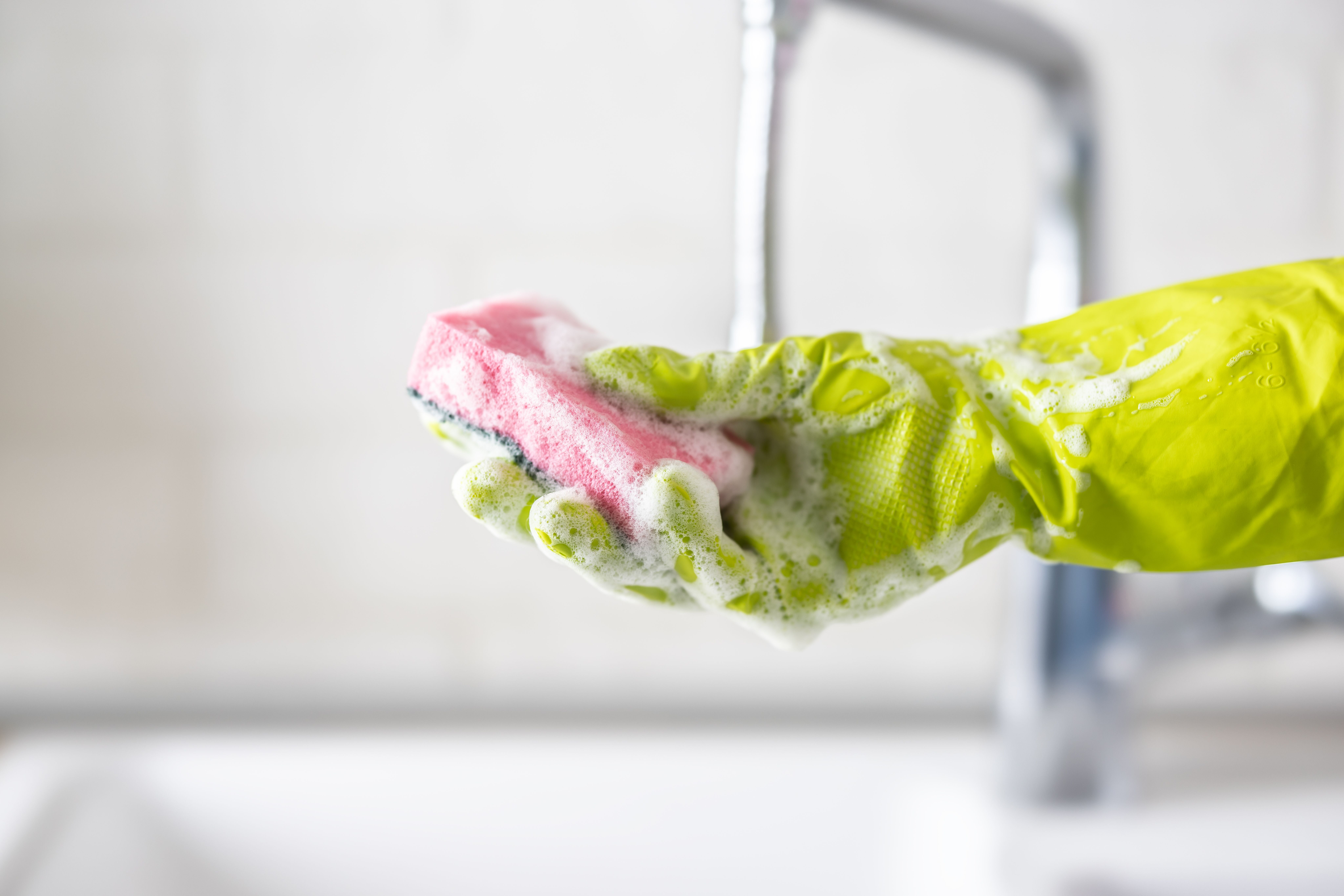 Frau hält einen schäumenden Schwamm mit gelbem Gummihandschuh über einem Waschbecken.