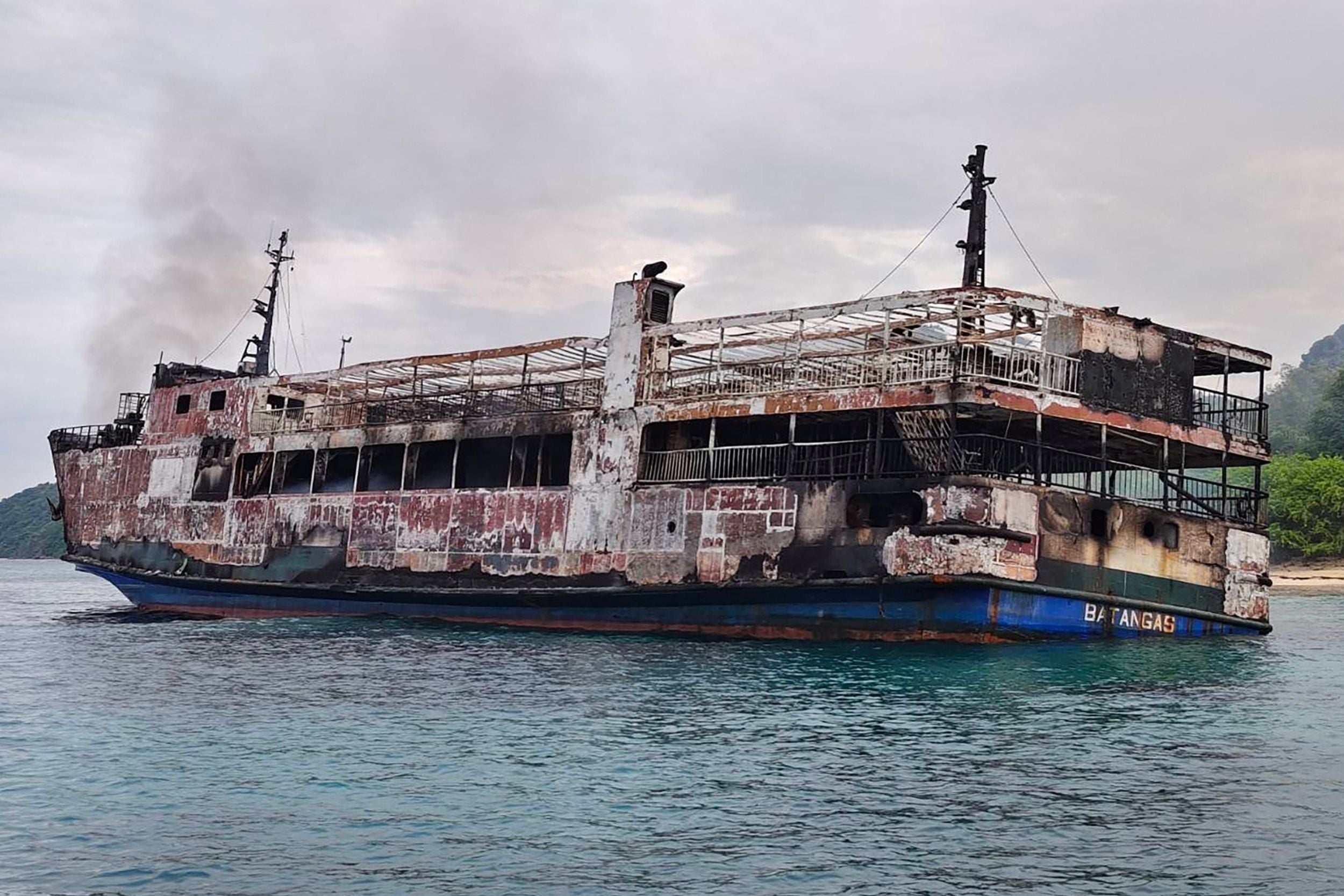 Ausgebranntes Fährschiff treibt beschädigt auf dem Meer nahe einer Küste bei bewölktem Himmel.