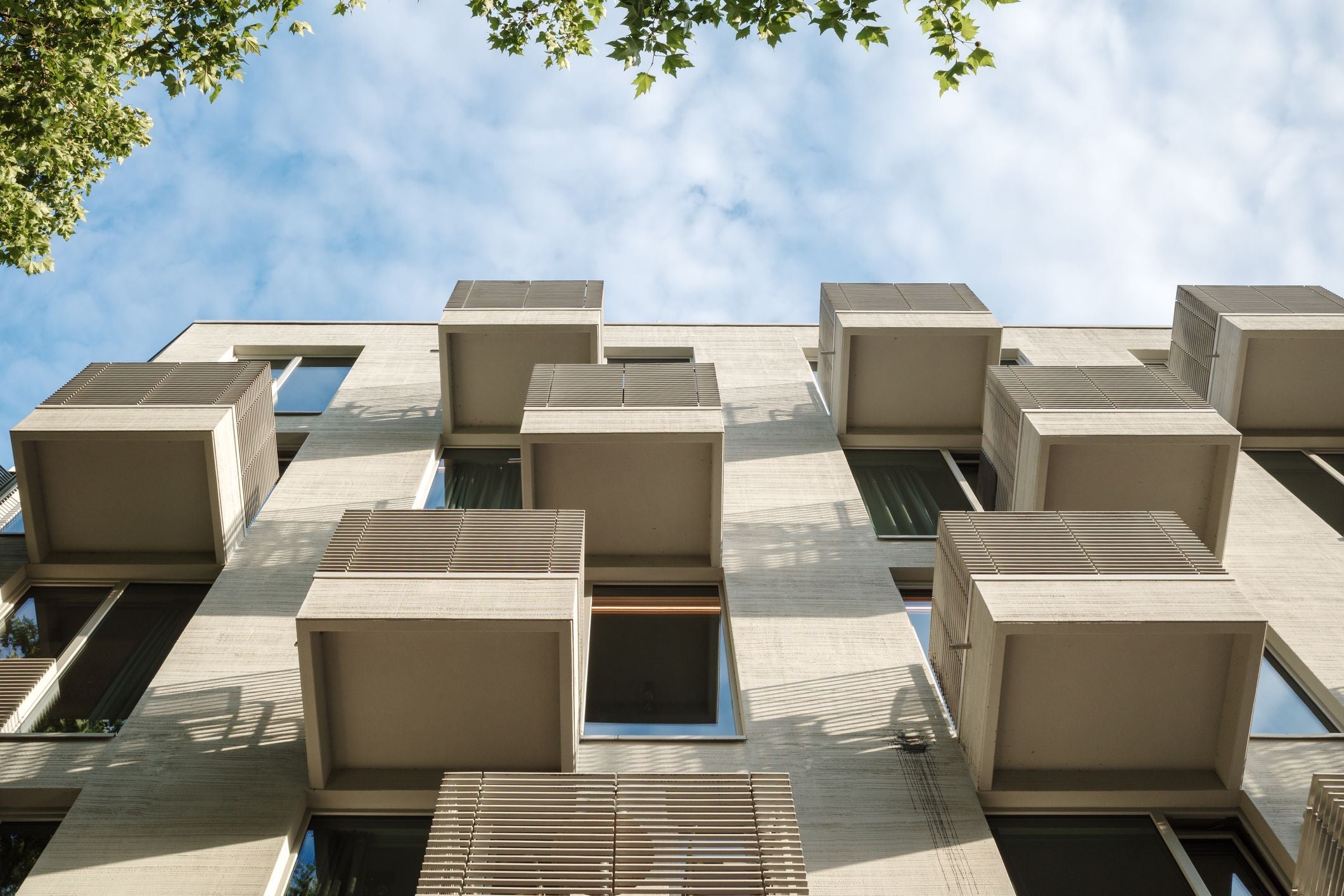 Moderne Wohnhausfassade mit hervorstehenden Box-Balkonen und blauem Himmel, von unten fotografiert.