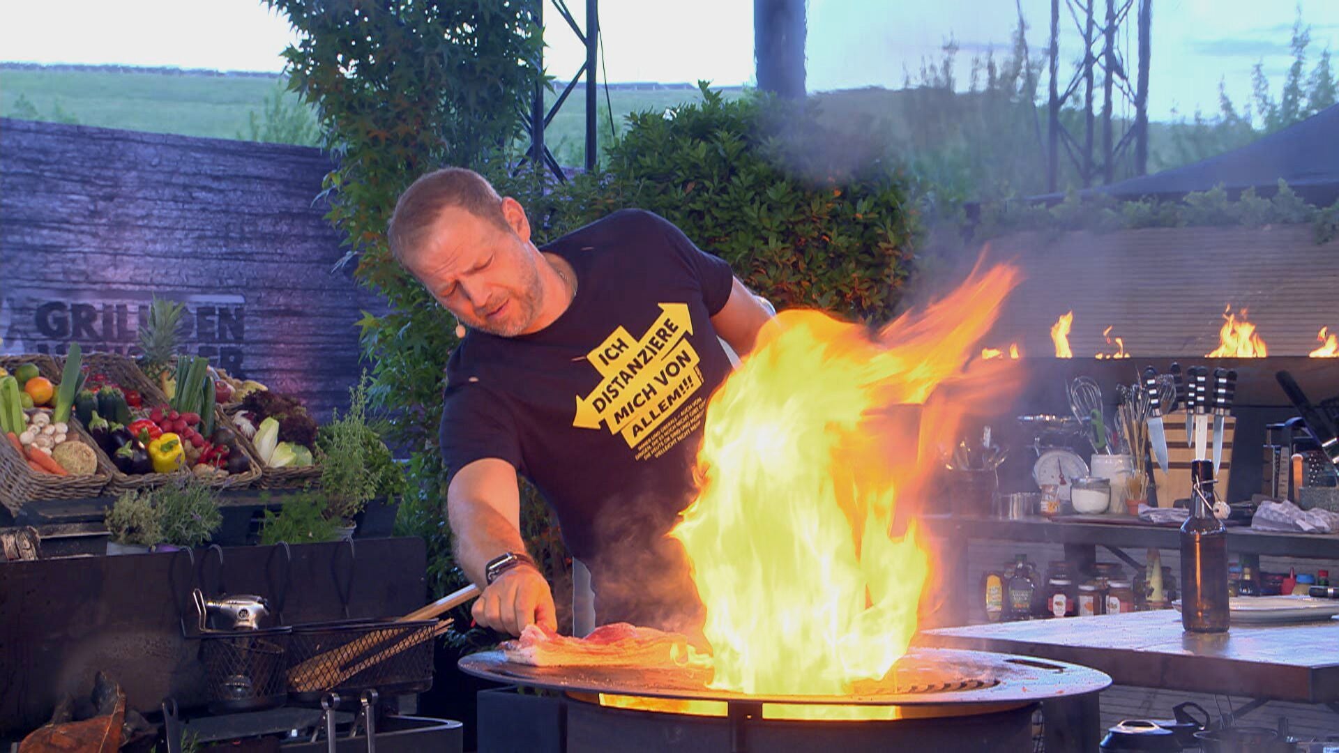 Ein Mann steht am Grill mit großer Flamme und kocht im Freien, Gemüse liegt im Korb daneben.