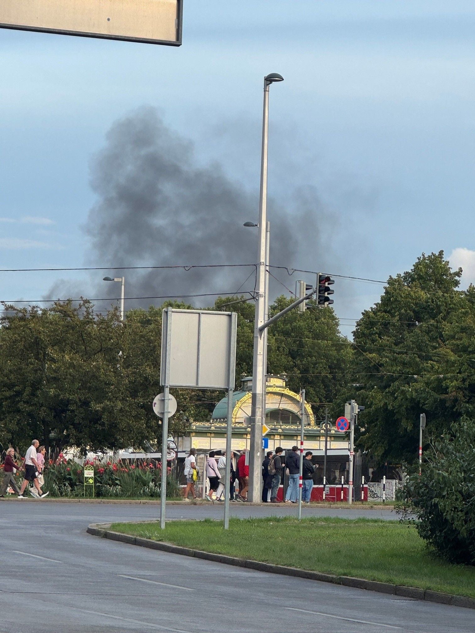 Schwarzer Rauch steigt hinter Bäumen am Karlsplatz auf, Passanten überqueren die Straße.
